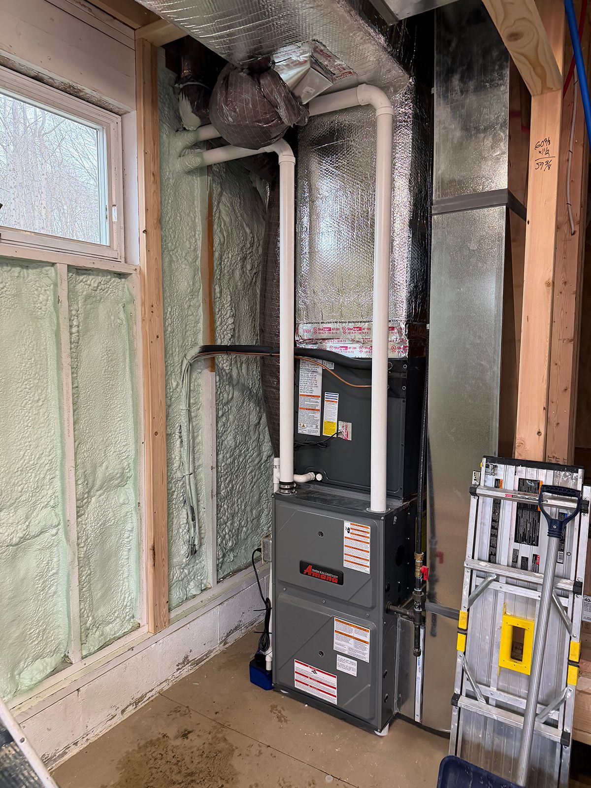 A grey high-efficiency gas furnace with white PVC venting pipes stands against a spray-foamed wall next to metal ductwork.