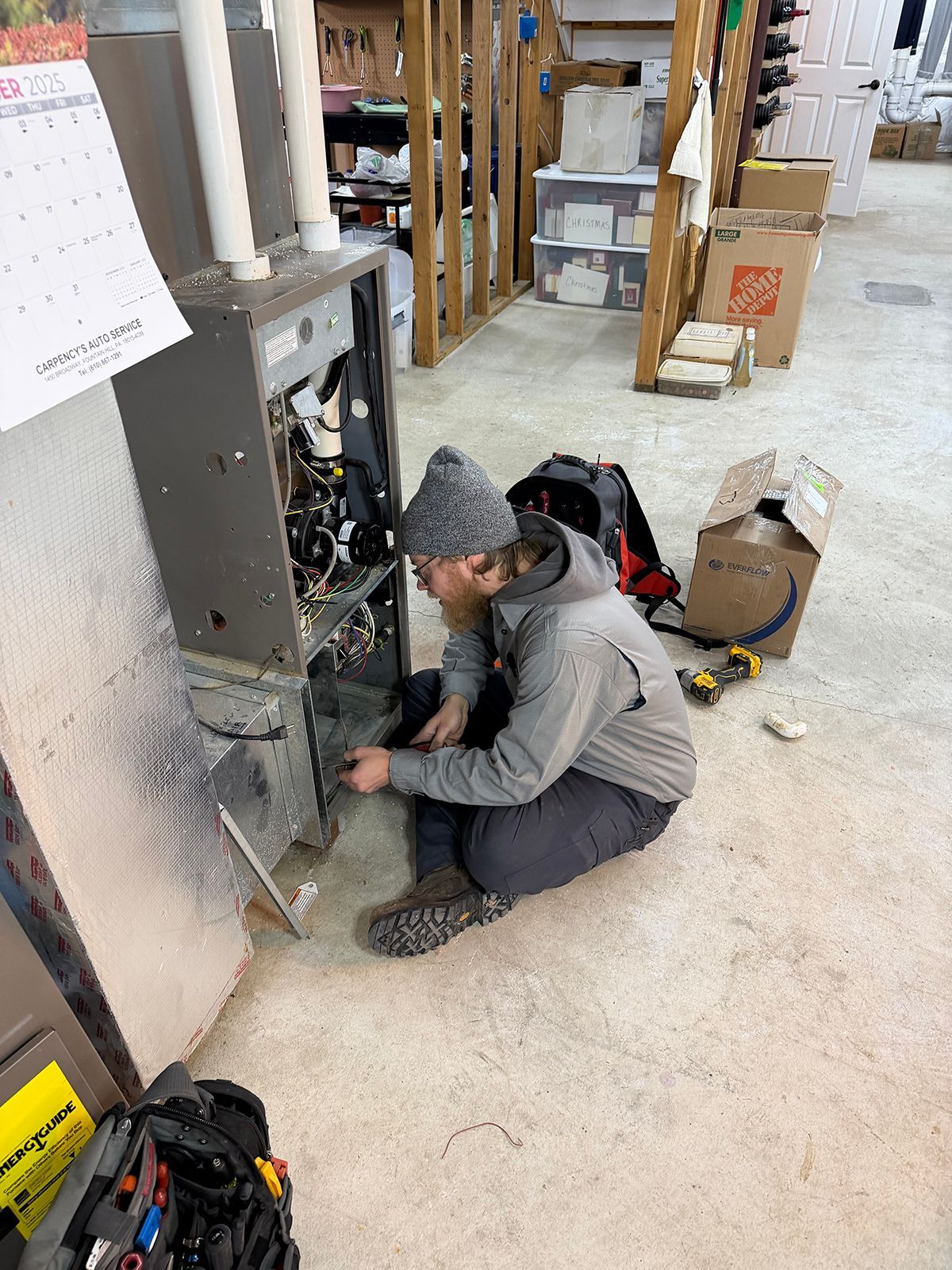 A technician wearing a grey hoodie and beanie sits on a concrete floor, repairing the components of an open furnace.