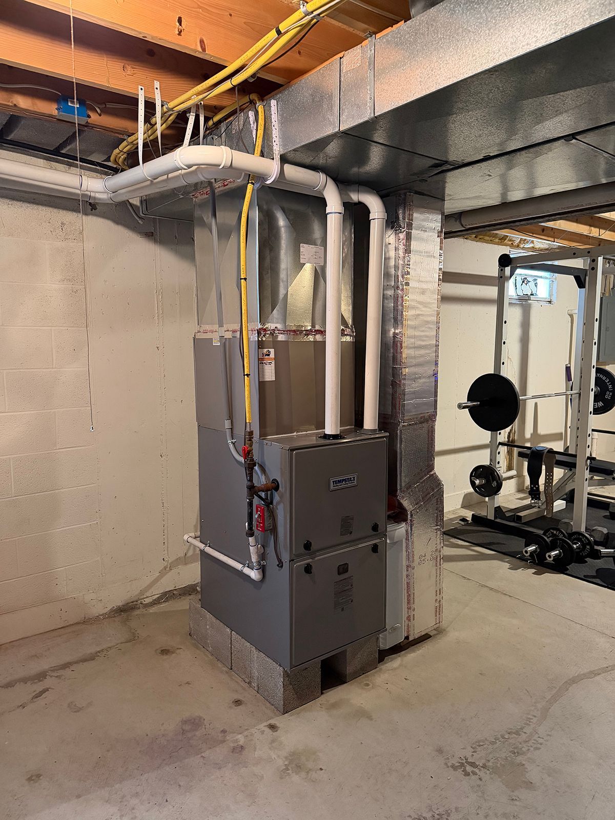 A tall, grey gas furnace with white PVC exhaust pipes sits on a concrete block in a basement next to a gym rack.