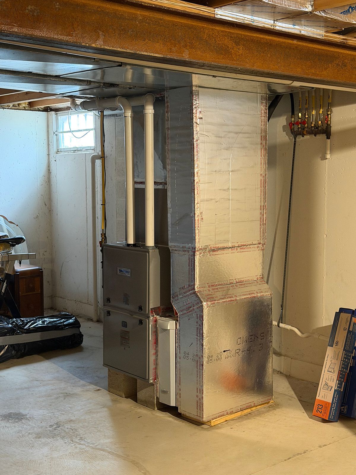A residential gas furnace and vertical silver metal ductwork installed in a concrete basement.