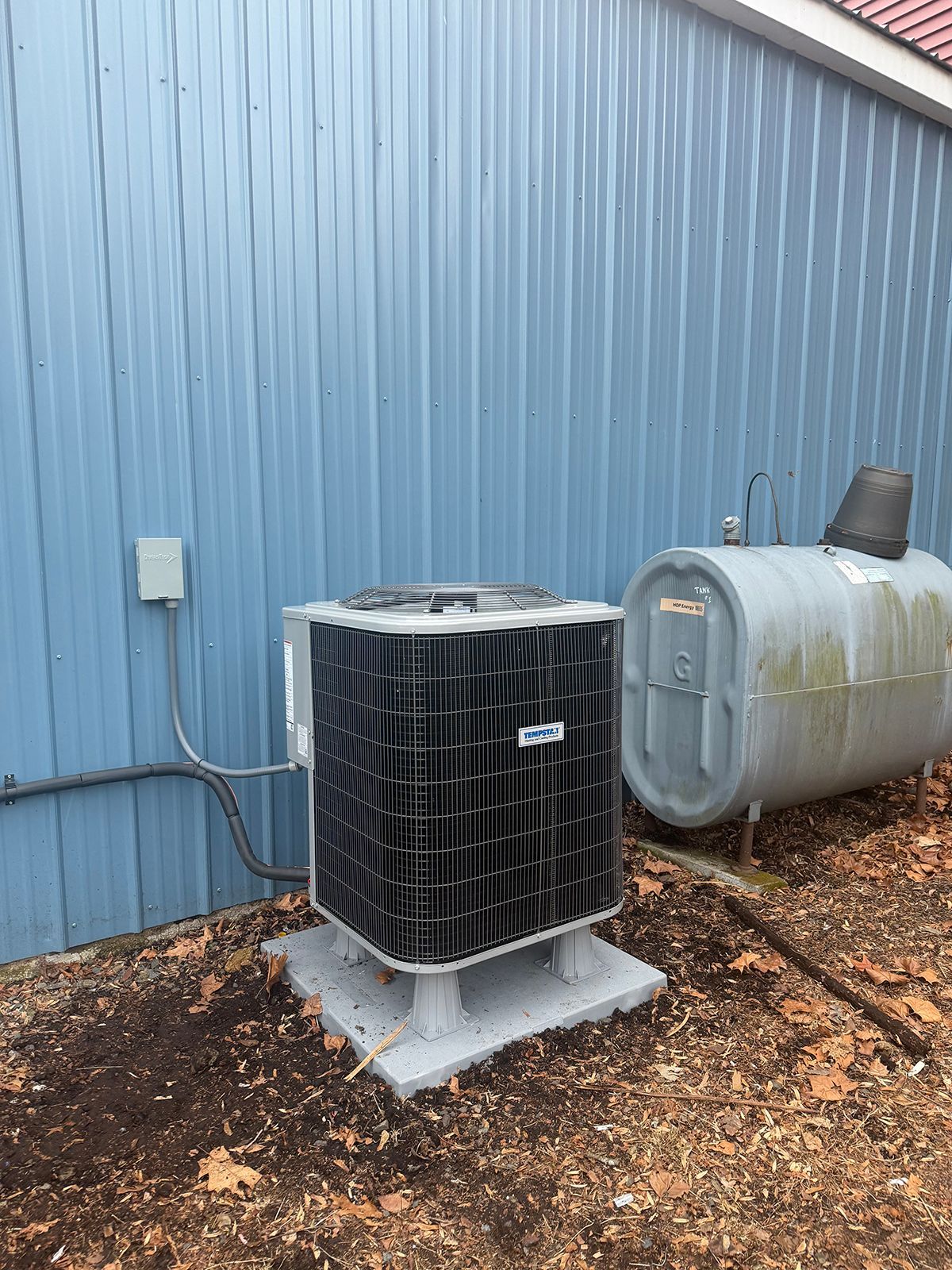 An outdoor air conditioner unit stands next to a cylindrical metal tank against a light blue metal-sided building.