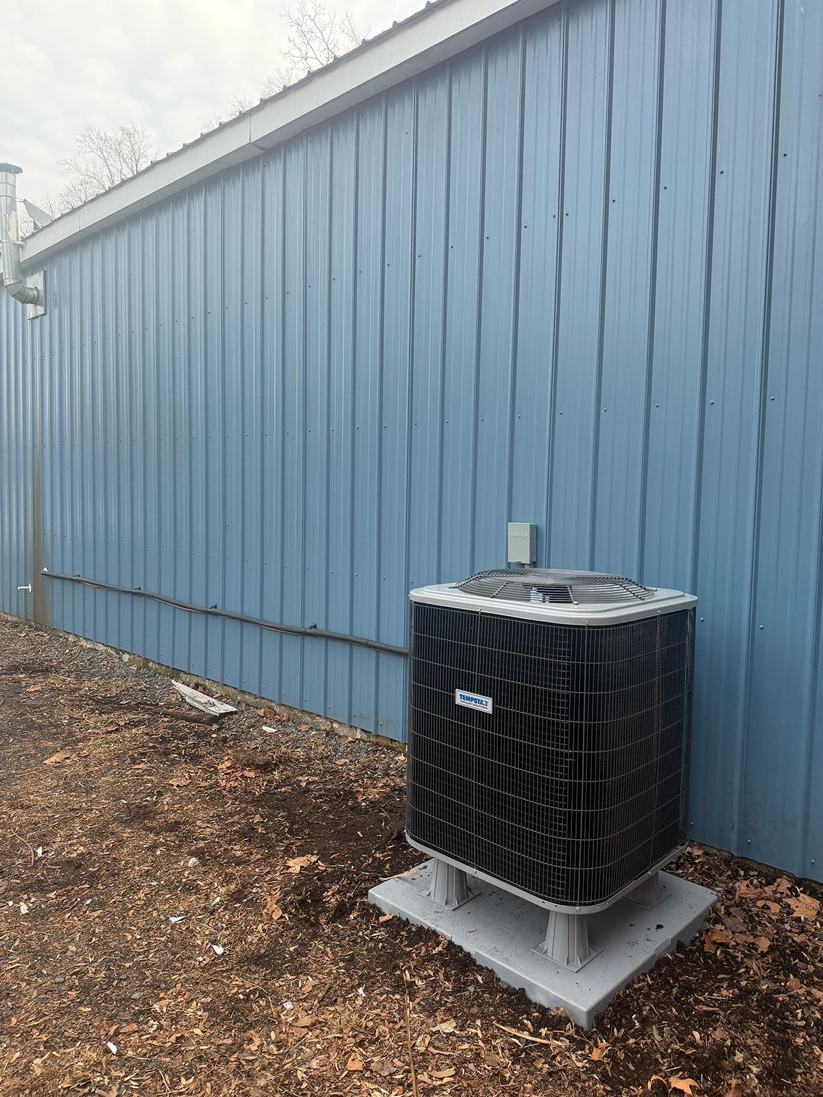 A central HVAC unit sits on a concrete pad against a blue corrugated metal wall on a bed of wood mulch.