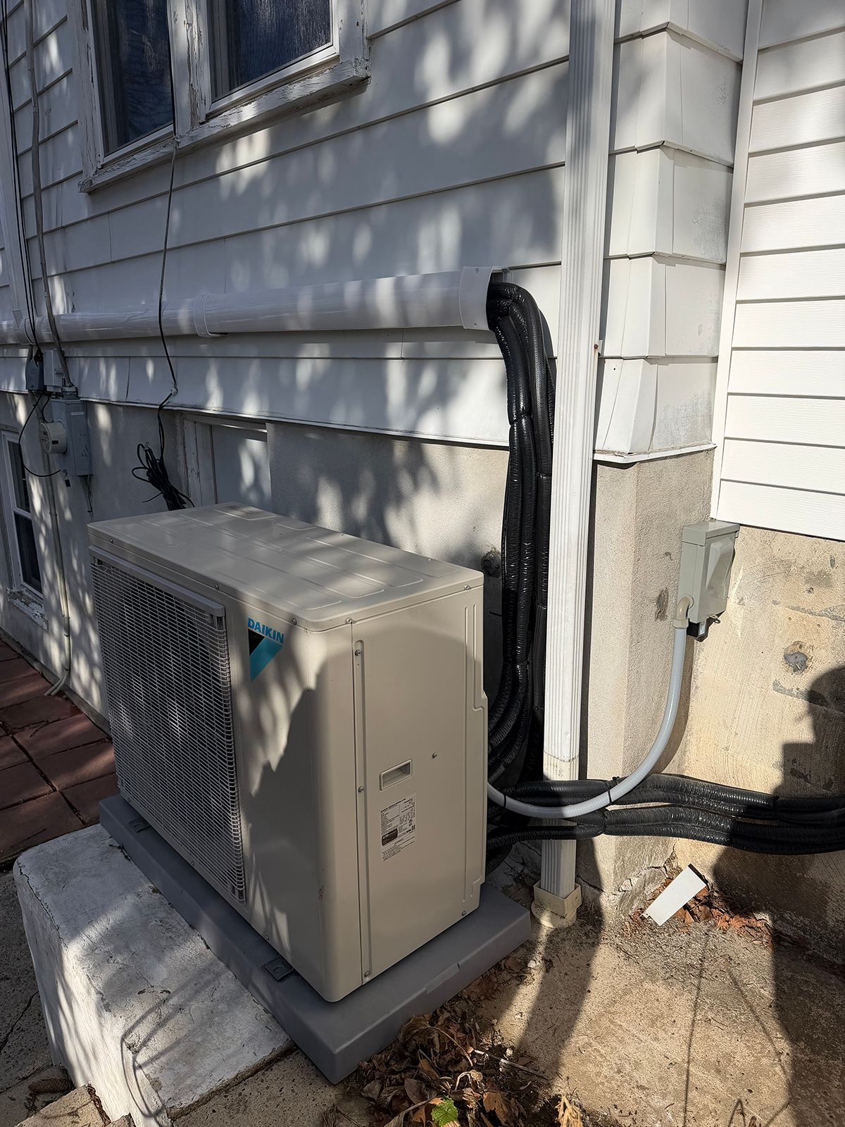 An outdoor heat pump unit sits on a base next to a white house with cables running up the exterior wall.