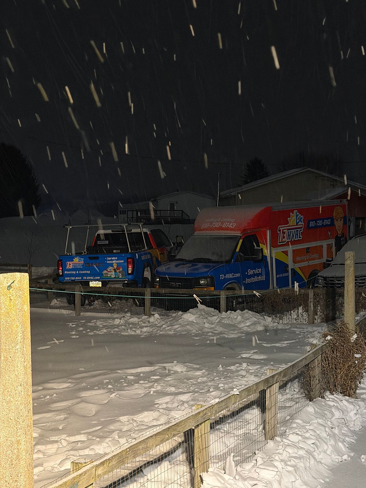 A blue pickup truck and a bright orange and blue delivery van parked in a snow-covered lot during a nighttime snowfall.