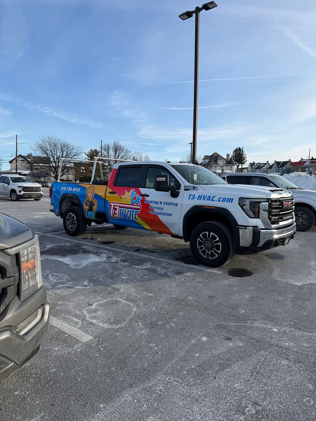 A white GMC pickup truck with colorful promotional graphics, parked in a sunny, snowy outdoor parking lot.
