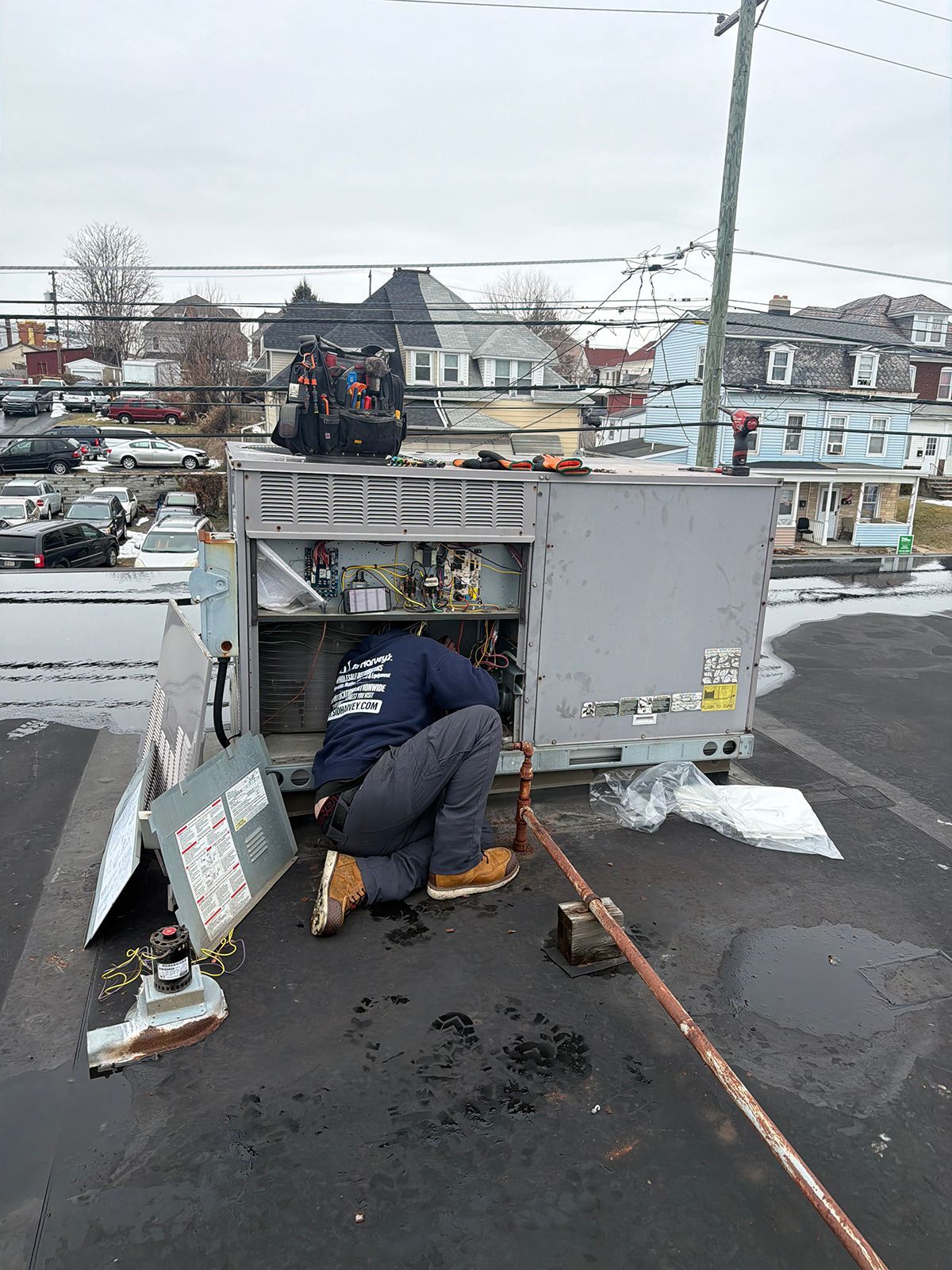 A technician in a blue uniform kneeling on a flat roof, working on the exposed electrical components of an HVAC unit.