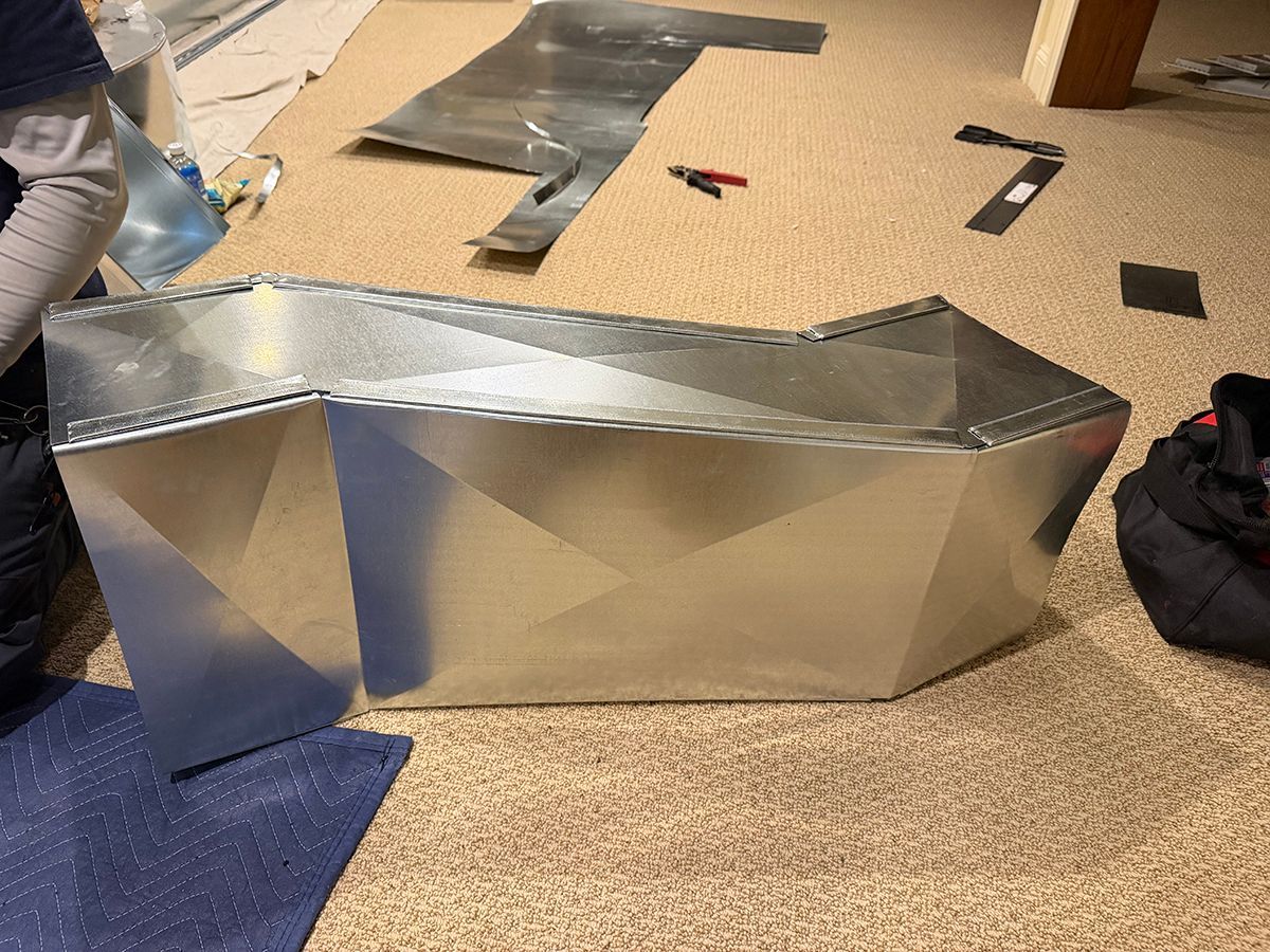A person fabricates a custom sheet metal duct piece on a carpeted floor, surrounded by tools and metal scraps.