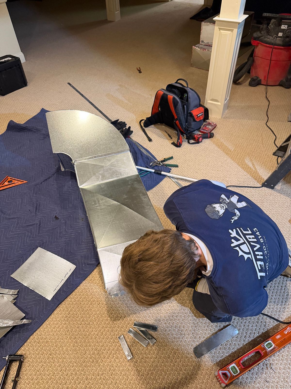 A technician in a blue uniform works on a metal HVAC duct piece on a carpeted floor, surrounded by various hand tools.