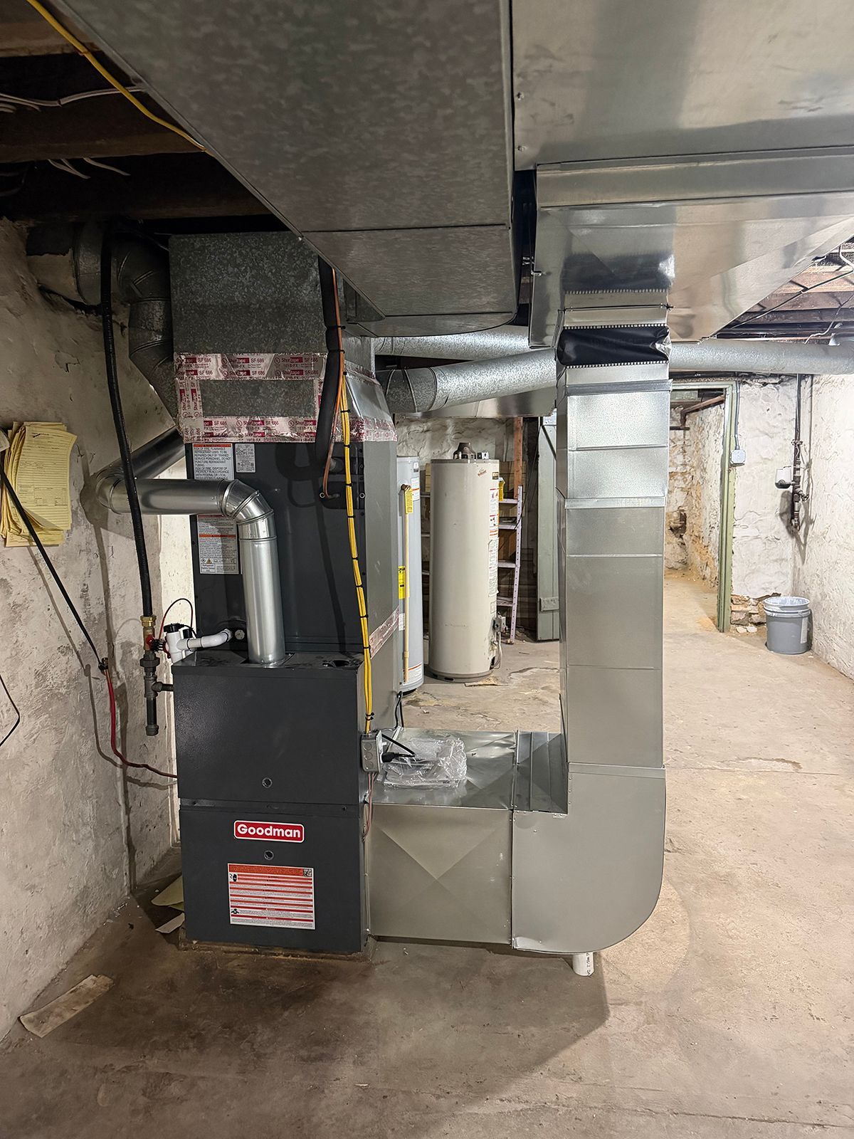 A Payne furnace unit connected to silver metal ductwork in an unfinished basement with a water heater in the background.