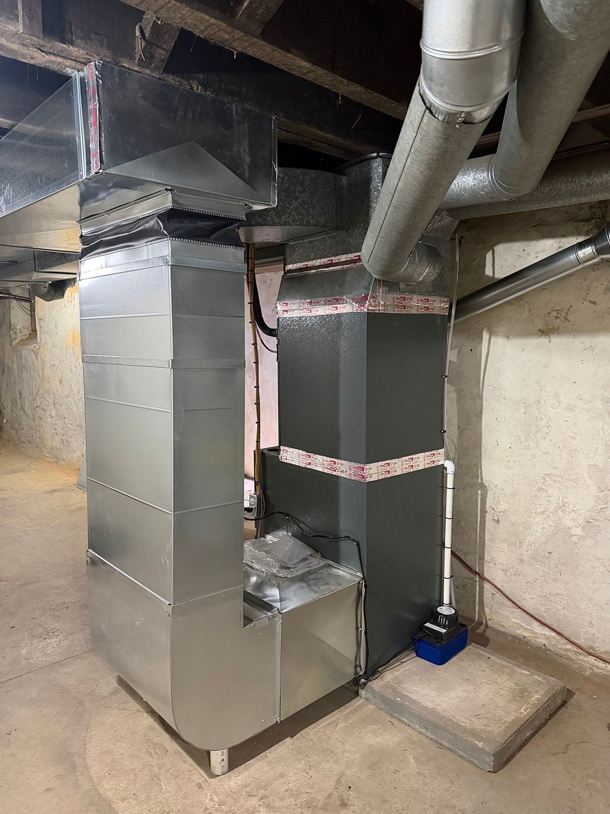 A new HVAC air handler with silver metal ductwork, a white condensation line, and a blue condensate pump in a basement.