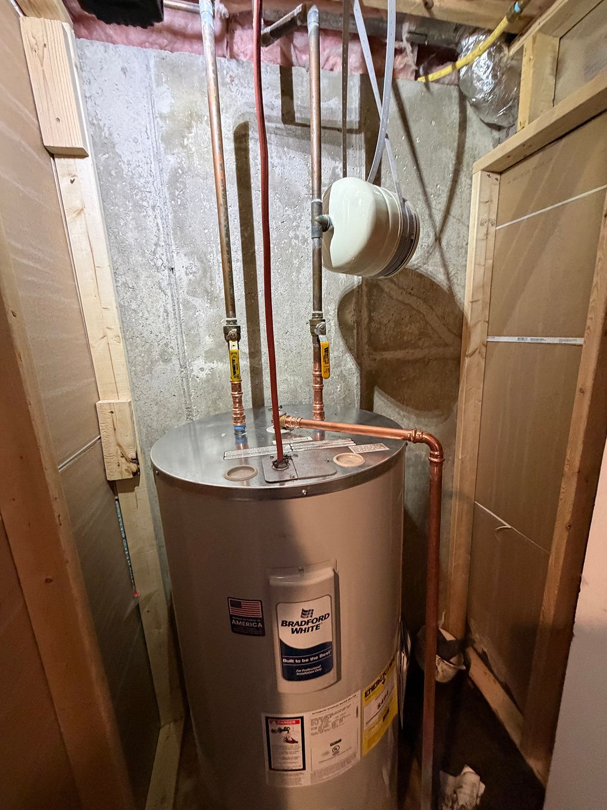 A water heater in a framed basement nook with connected copper pipes and a small white expansion tank.