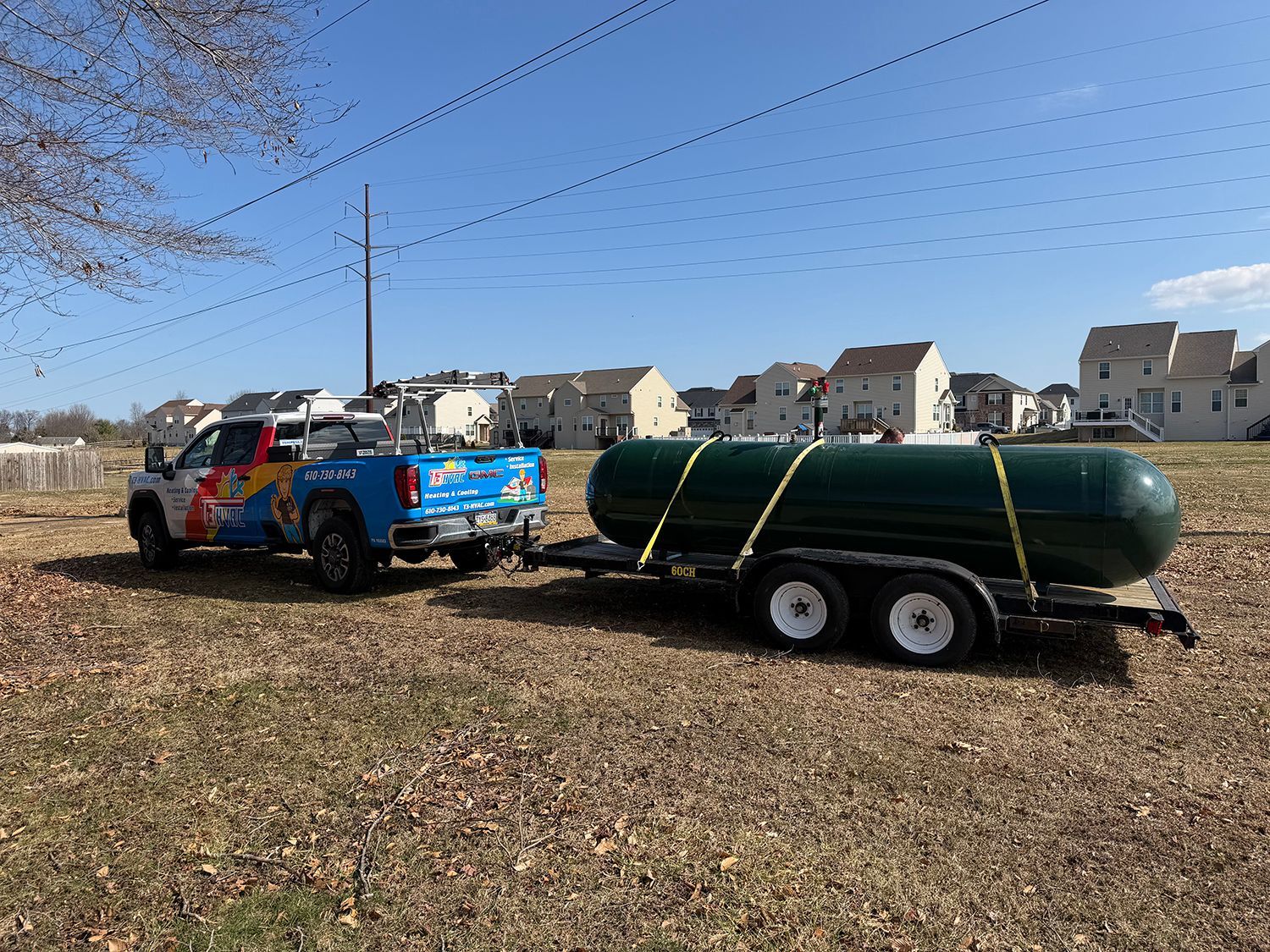 A blue pickup truck towing a large, dark green propane tank on a trailer across a grassy field near suburban homes.