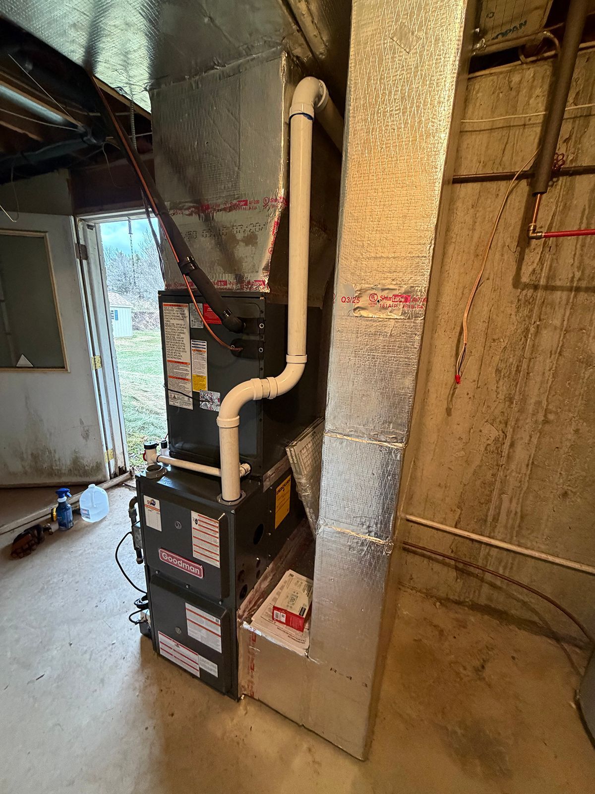 A furnace unit with connected metal ductwork and a white PVC drain pipe, located in a basement near an open door.