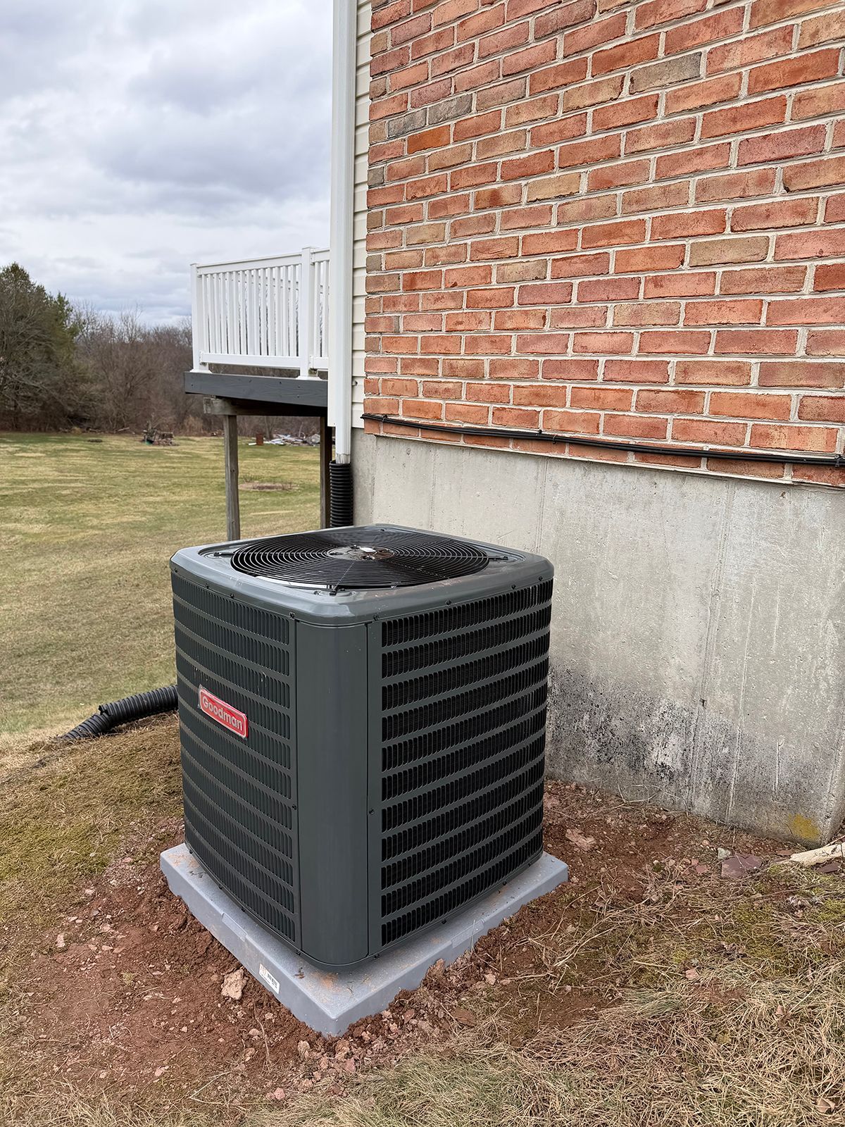 A dark gray HVAC unit sits on a concrete pad next to a brick house exterior under a cloudy sky.