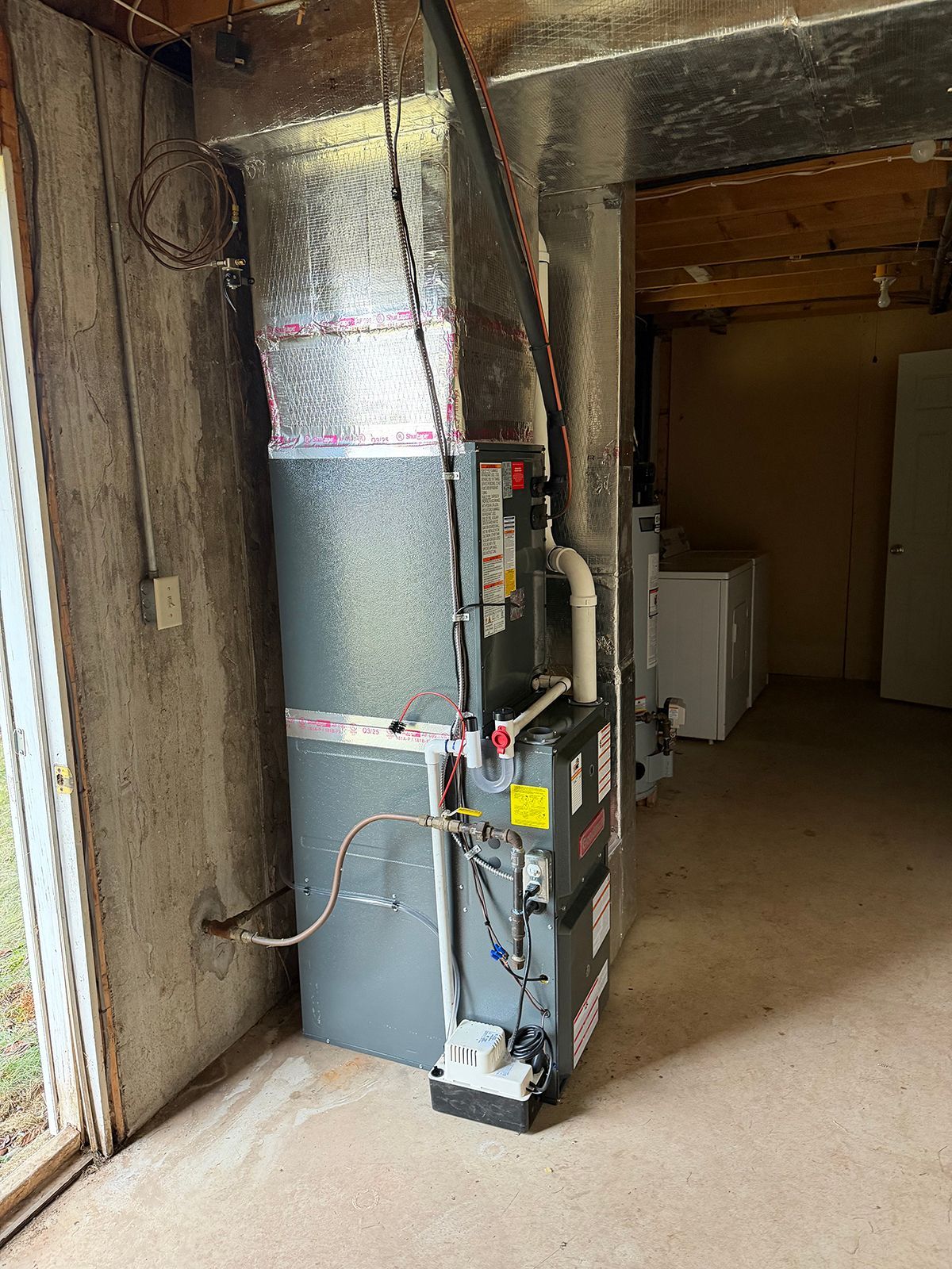 A gray HVAC furnace unit installed against a concrete basement wall with silver insulated ductwork above.