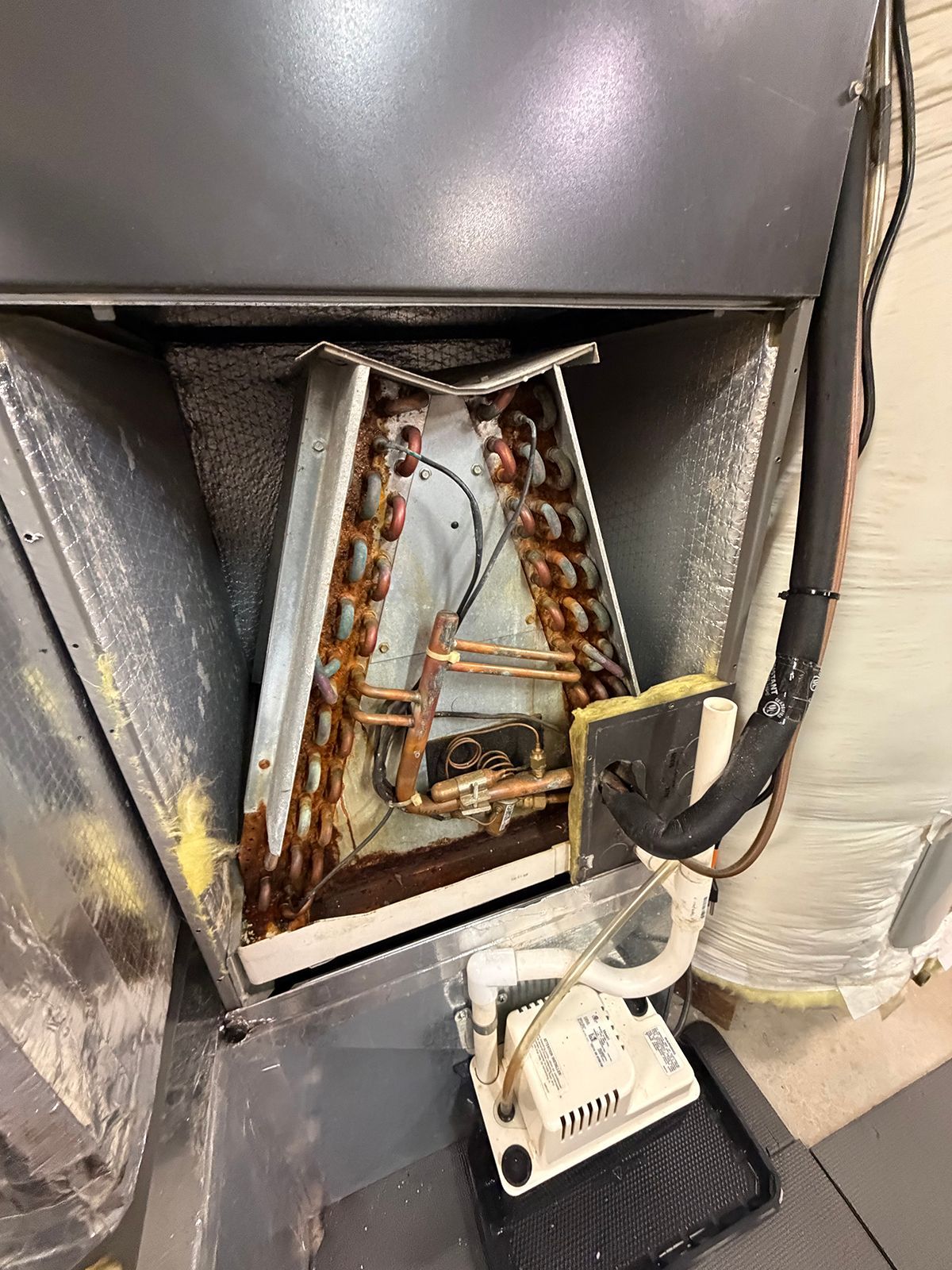 An open residential HVAC evaporator coil unit with copper tubing and an external condensate pump in a utility room.