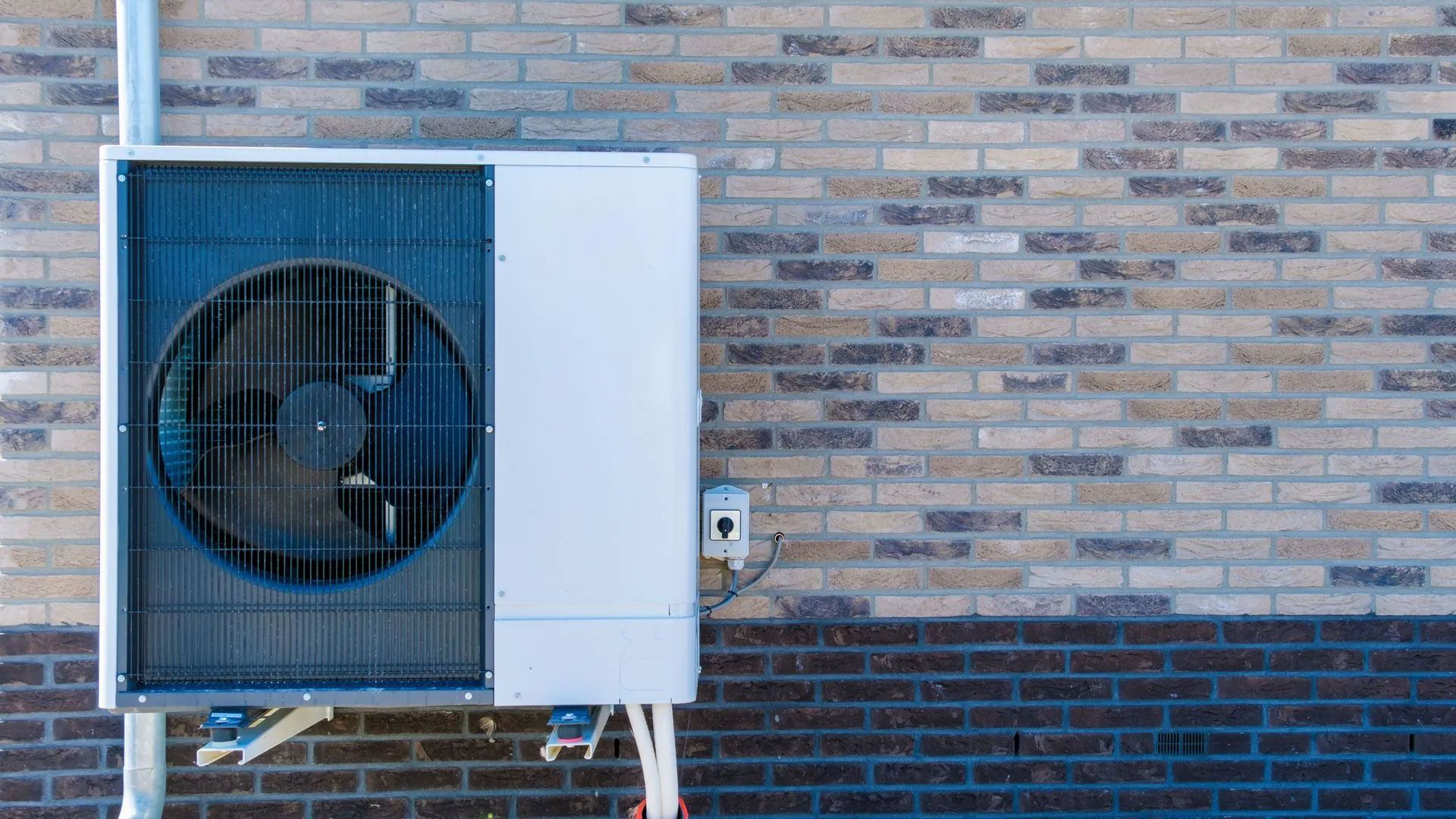 A white outdoor heat pump unit mounted on a brown and beige brick wall.
