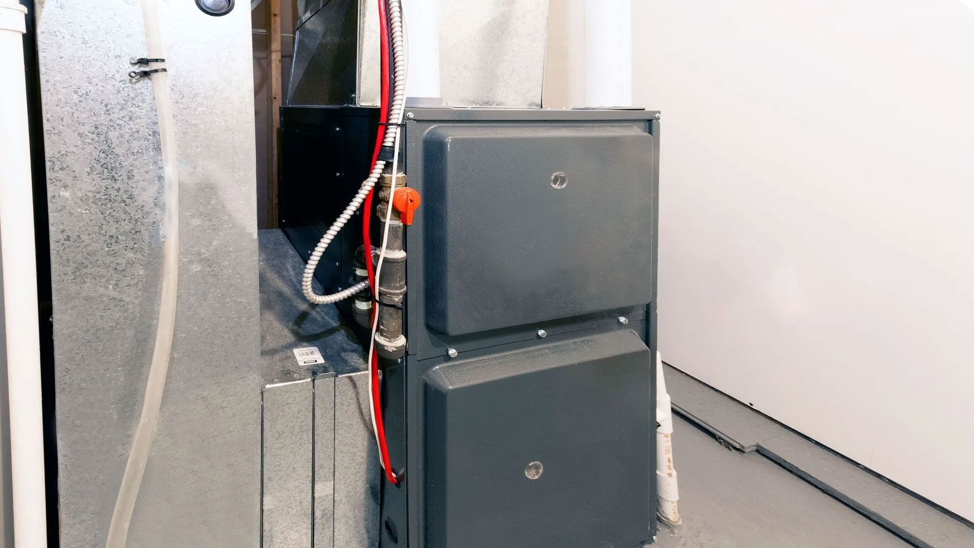 A gray residential furnace with a red gas shut-off valve and wiring attached to the side, set in a utility room.