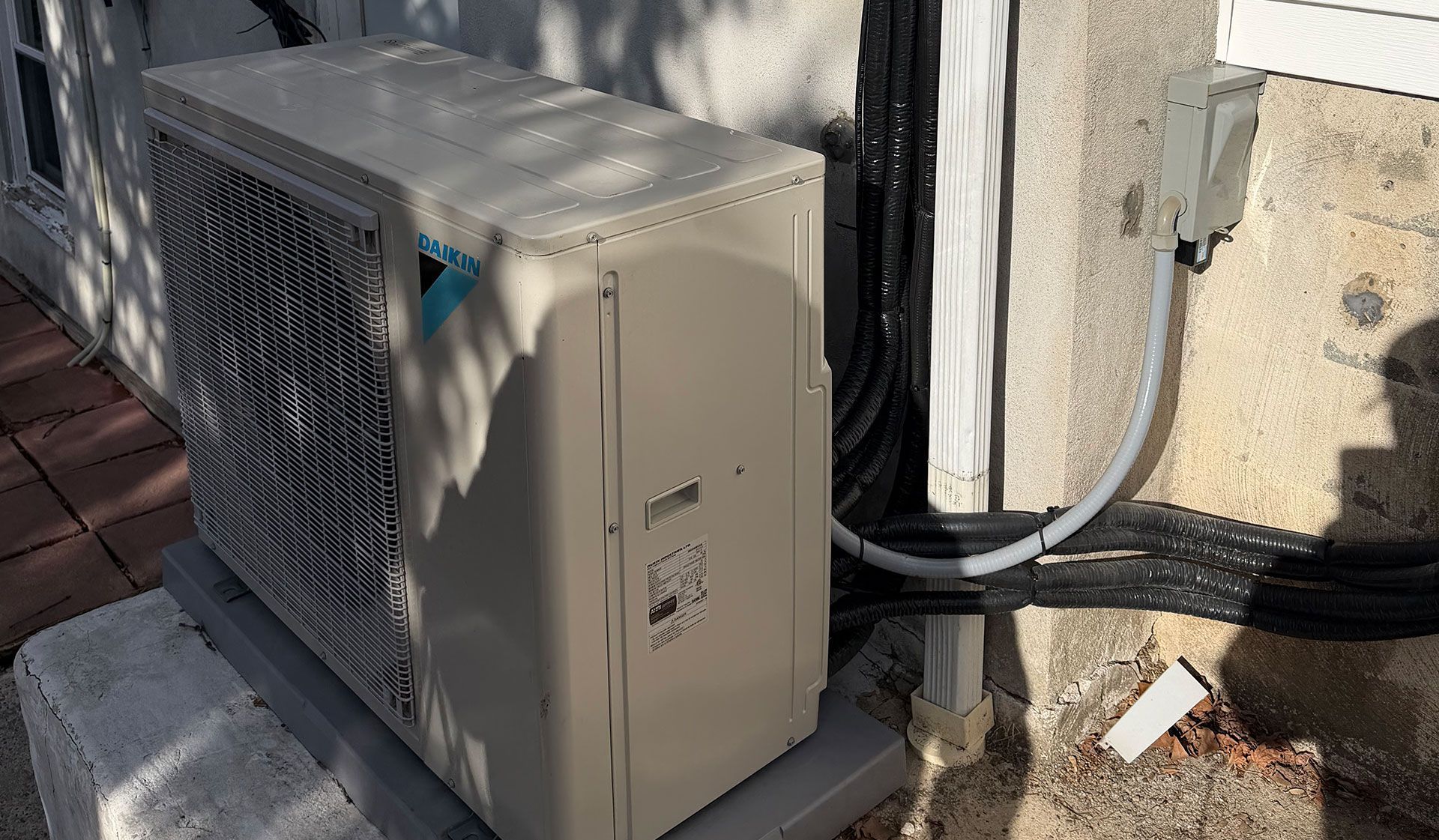 A beige Daikin outdoor HVAC condenser unit sits on a concrete pad next to a wall with black electrical conduits.