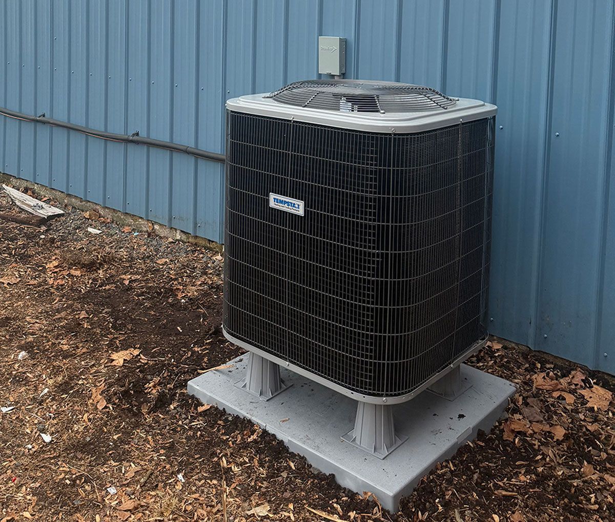 An outdoor central air conditioning unit sits on a gray concrete pad against a blue corrugated metal wall.