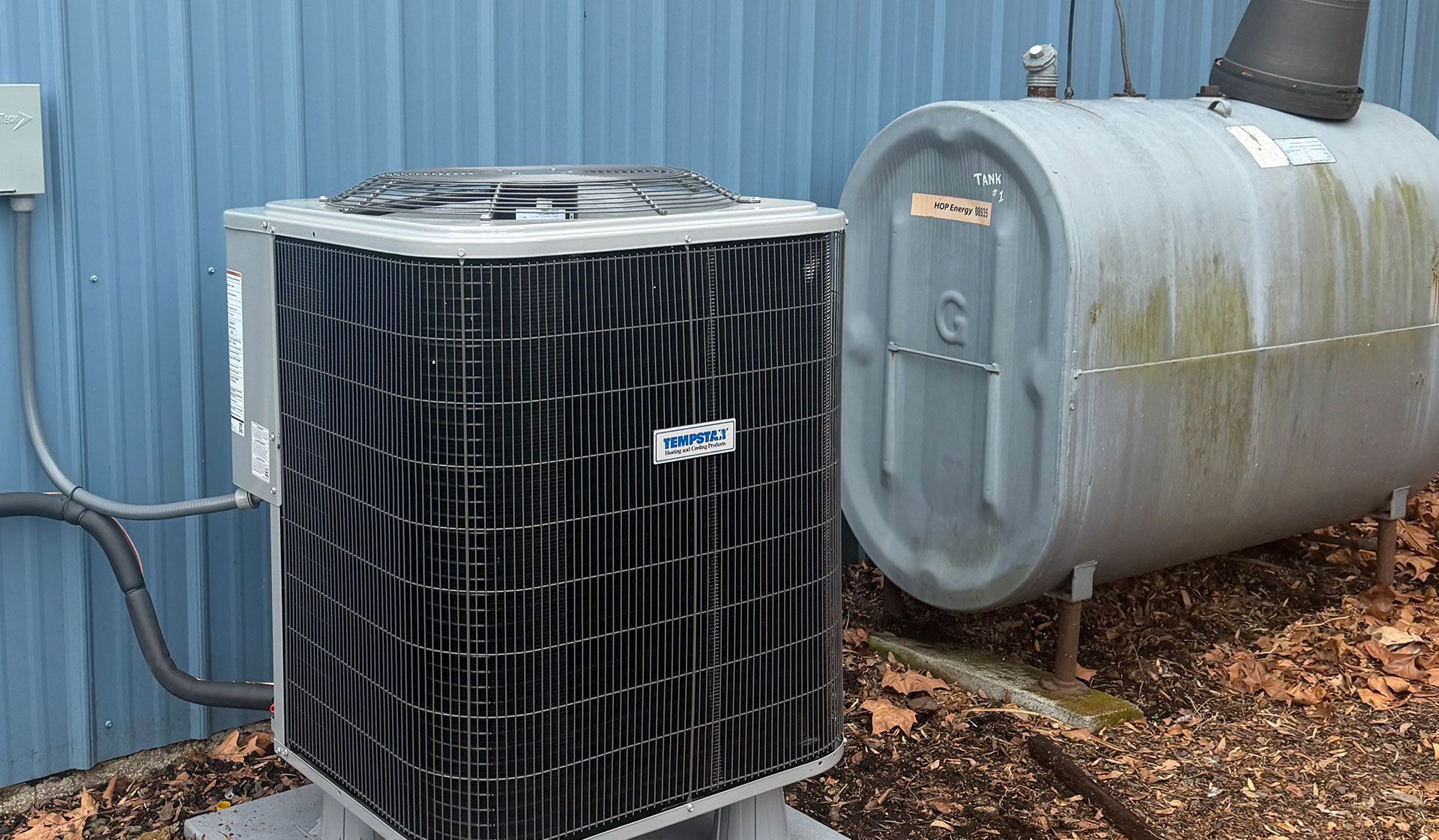 An outdoor air conditioning unit and a gray fuel oil storage tank sit on a bed of leaves against a blue metal wall.