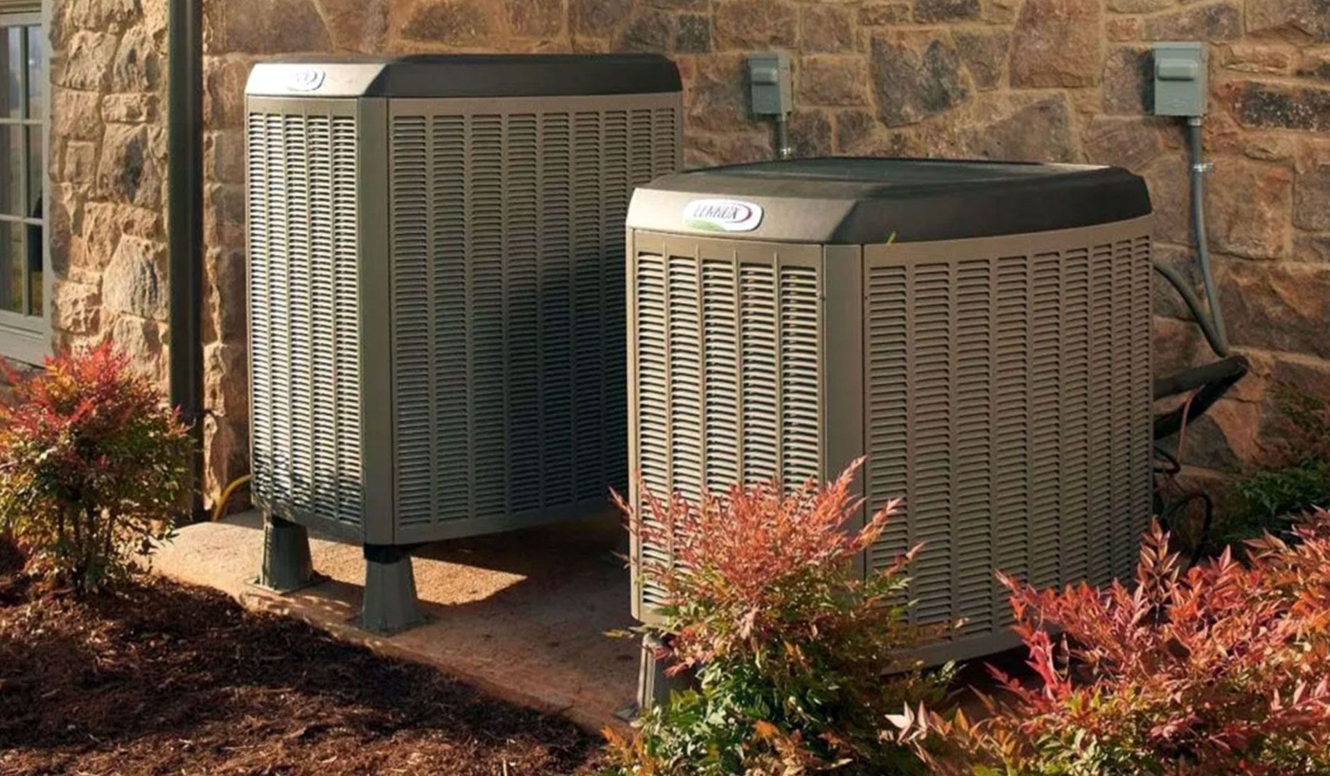 Two beige outdoor HVAC units sitting on a concrete pad against a stone exterior wall, surrounded by shrubs.