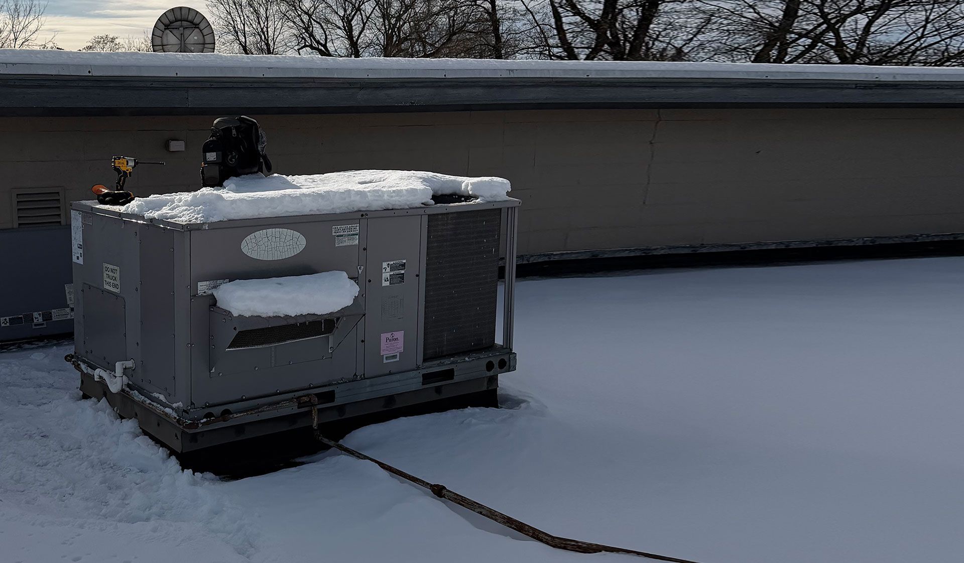 A rooftop HVAC unit covered in snow on a sunny winter day, with a utility cable extending across the snowy roof.