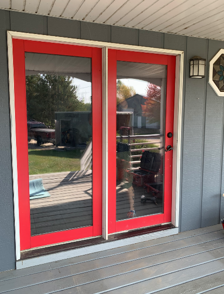 Red-framed glass double doors on a gray house porch reflecting the backyard.