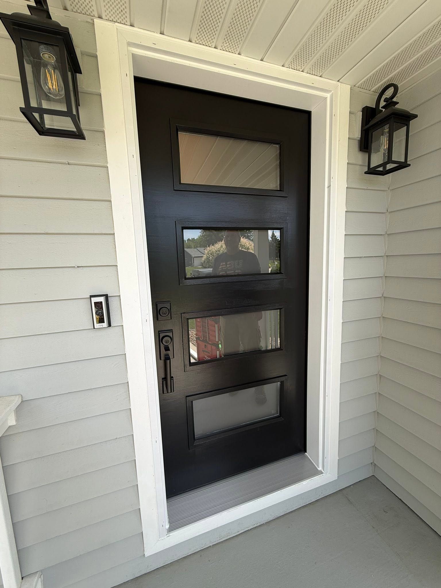 Black front door with three frosted glass panels, framed by white trim and flanked by wall lanterns.