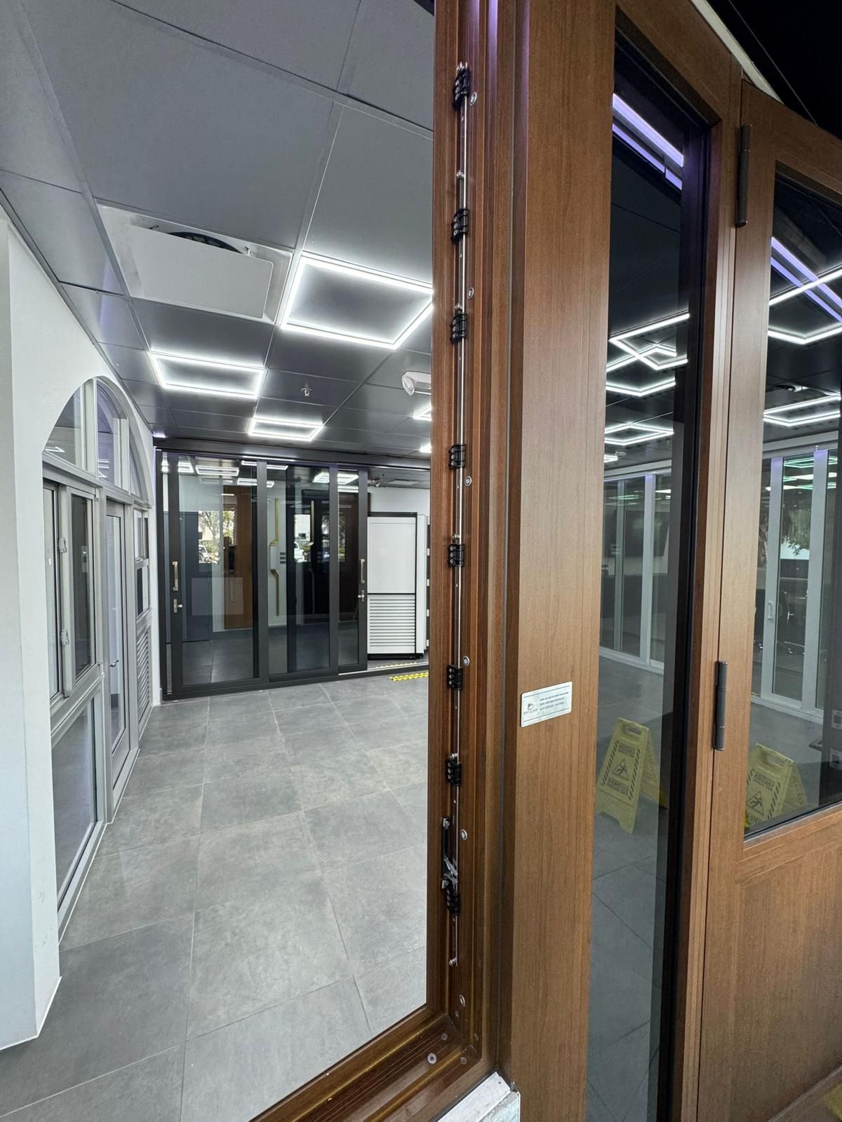 A view through a brown-framed window into a hallway. The interior has gray tile and recessed lighting.