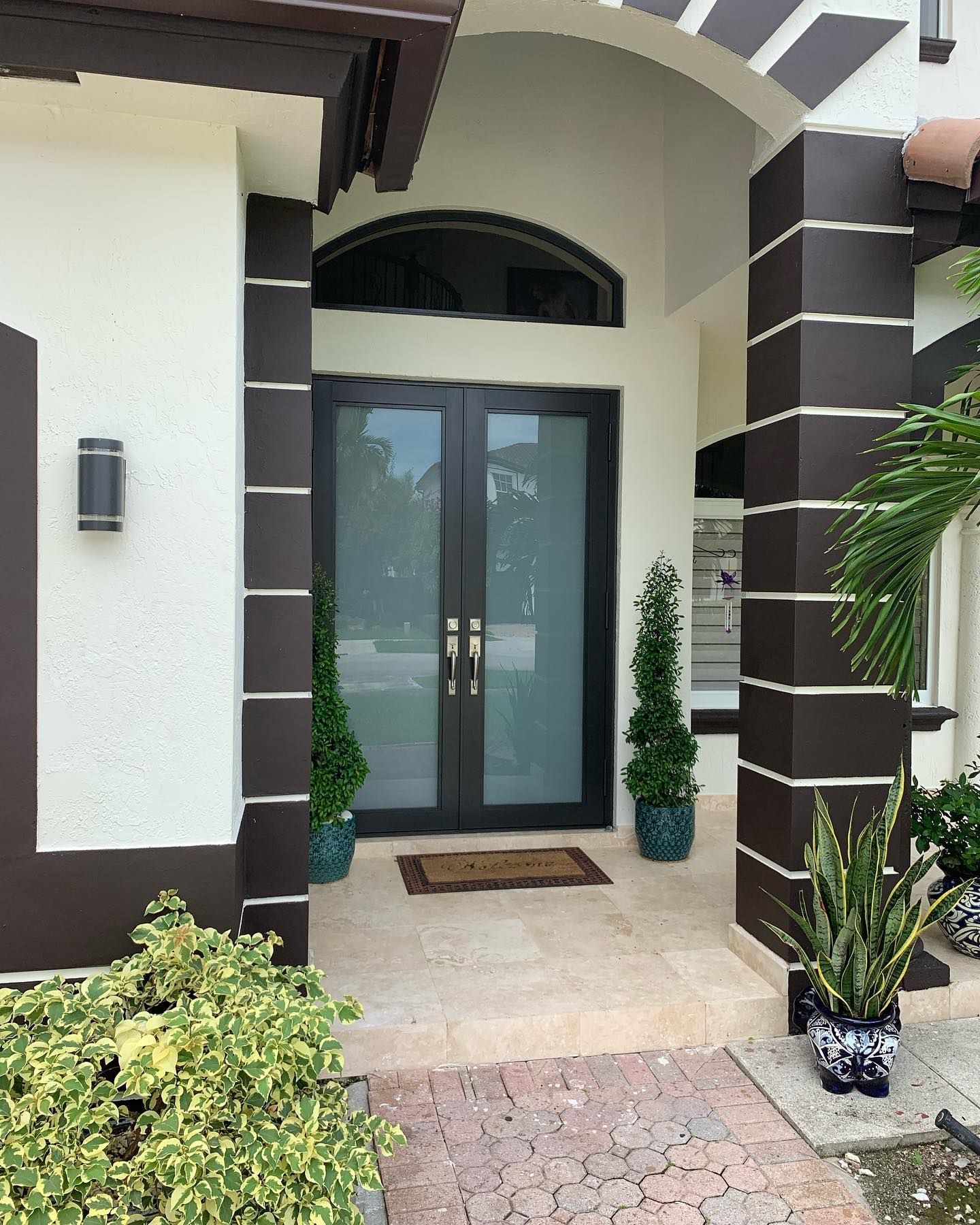 Front entrance of a house with double glass doors, columns, and a walkway. Dark brown and white exterior with potted plants.