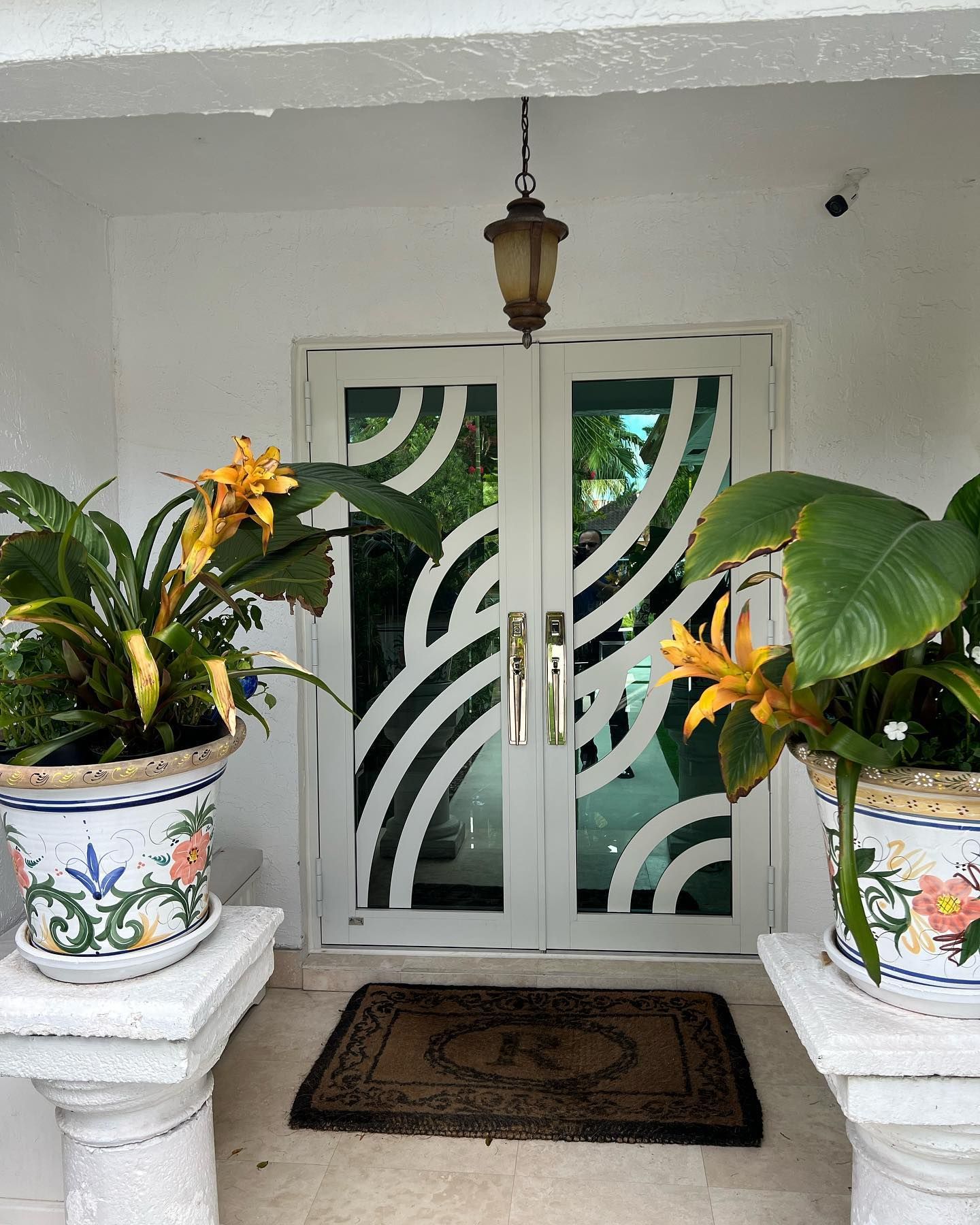 White arched entry doors with curved glass design, flanked by potted plants on pedestals.