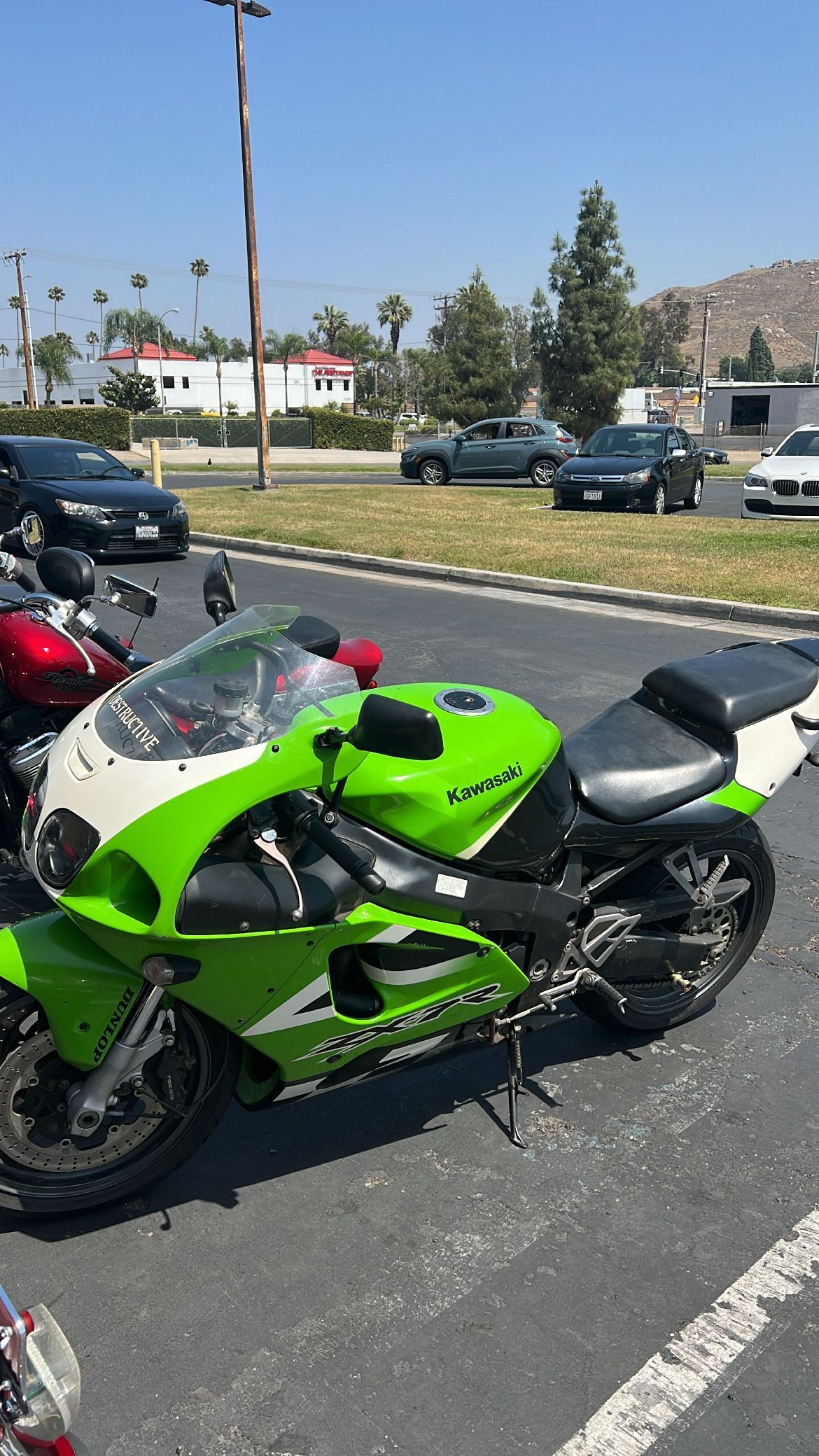 Green and white Kawasaki sport bike parked in a lot, with cars and buildings in the background on a sunny day.