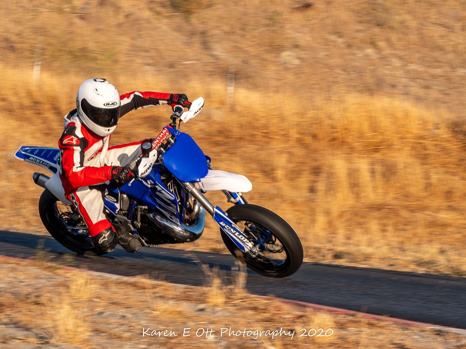 Motorcyclist on blue and white bike racing on dirt road; rider in red and white suit.
