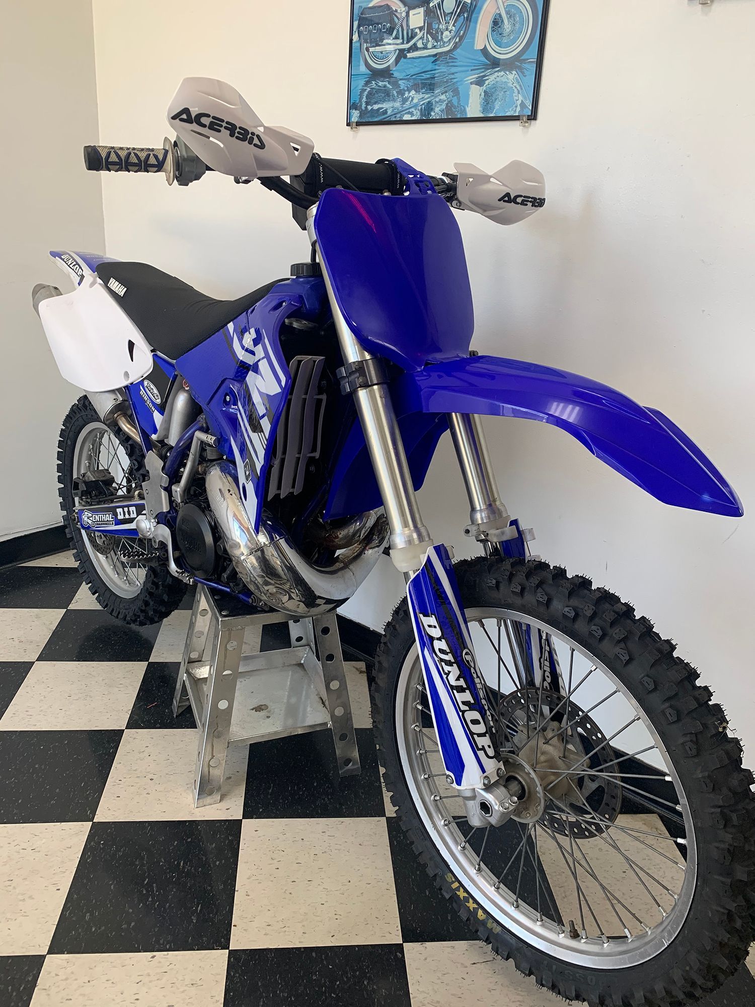 Blue Yamaha dirt bike on a stand, indoors with a checkered floor and a poster on the wall.