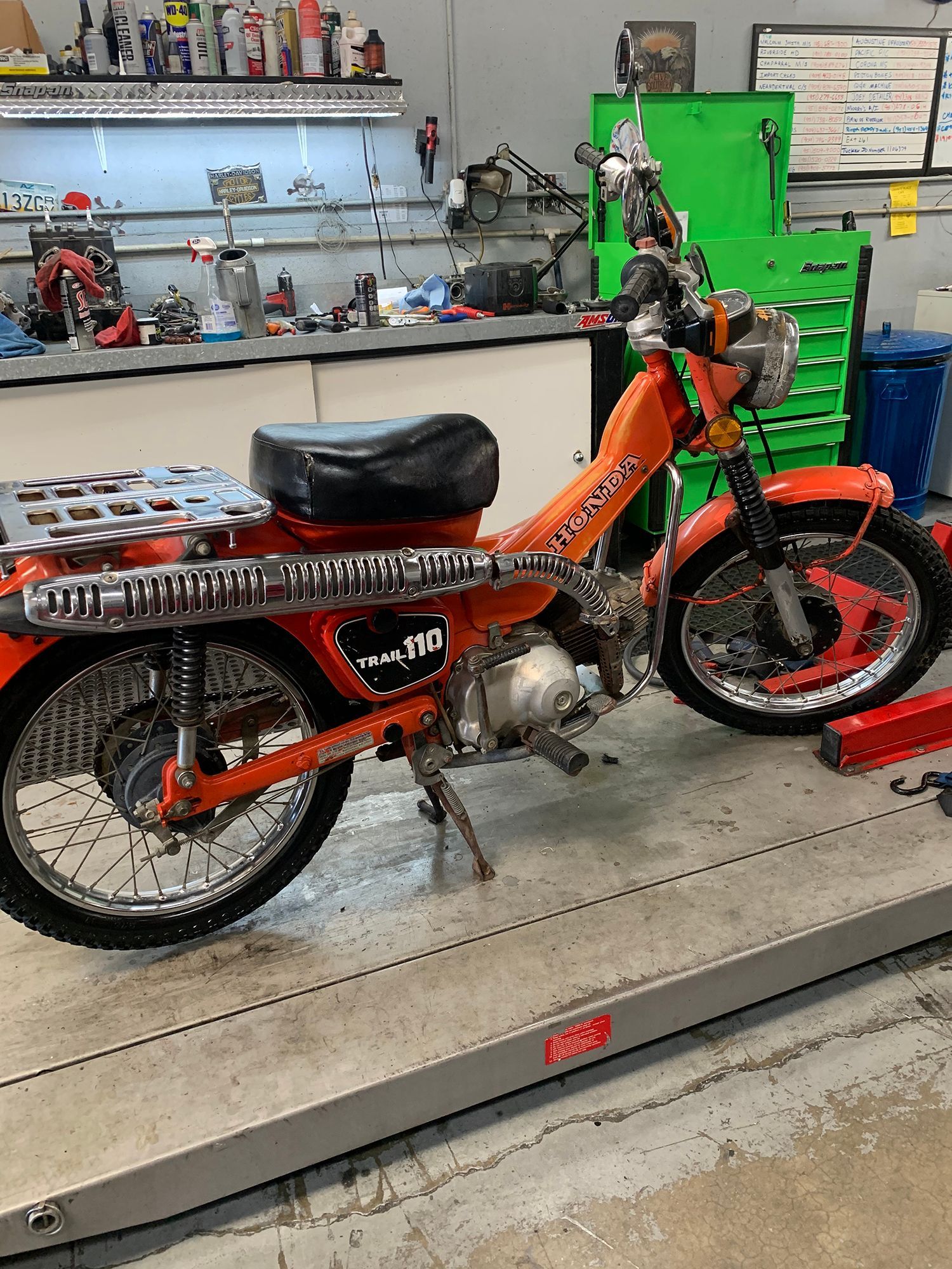 Orange Honda Trail 90 motorcycle in a workshop, featuring a silver rack, black seat, and bright orange frame.