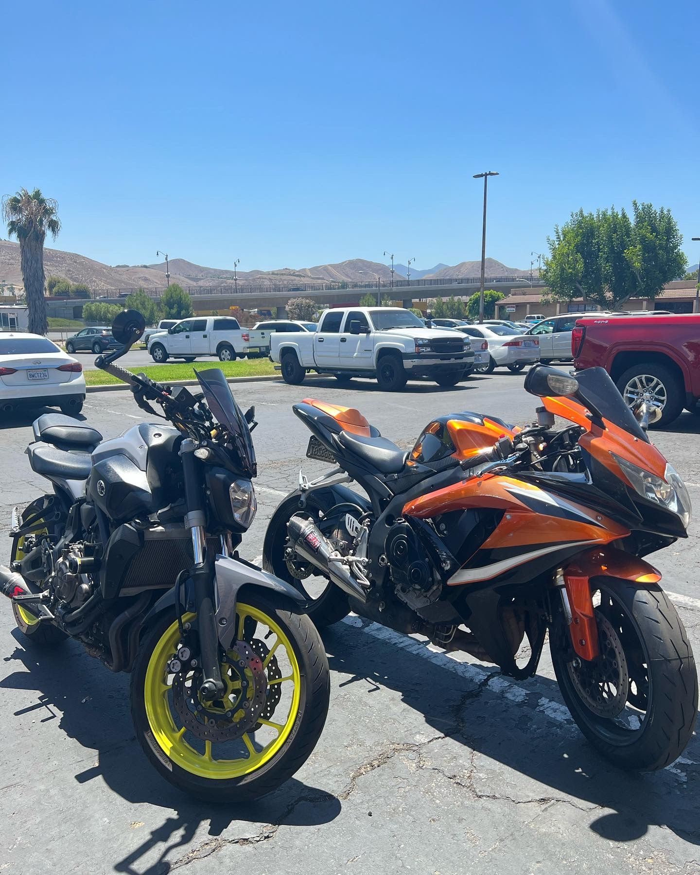 Two motorcycles parked outside on a sunny day. One is grey with yellow wheels, the other is orange. Cars are in the background.