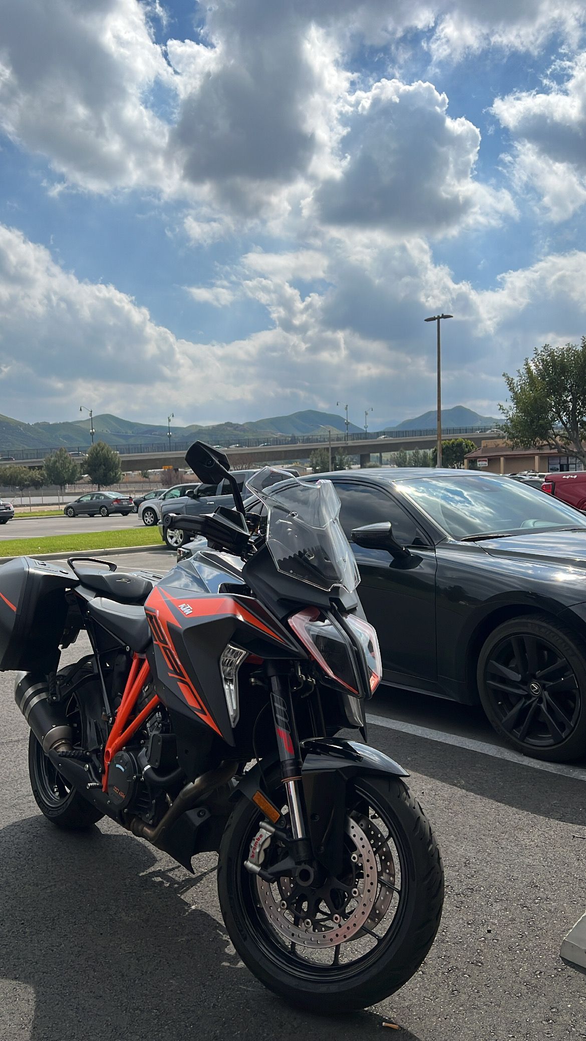 Black and orange motorcycle parked next to a black car, under a cloudy sky. Mountains in the background.