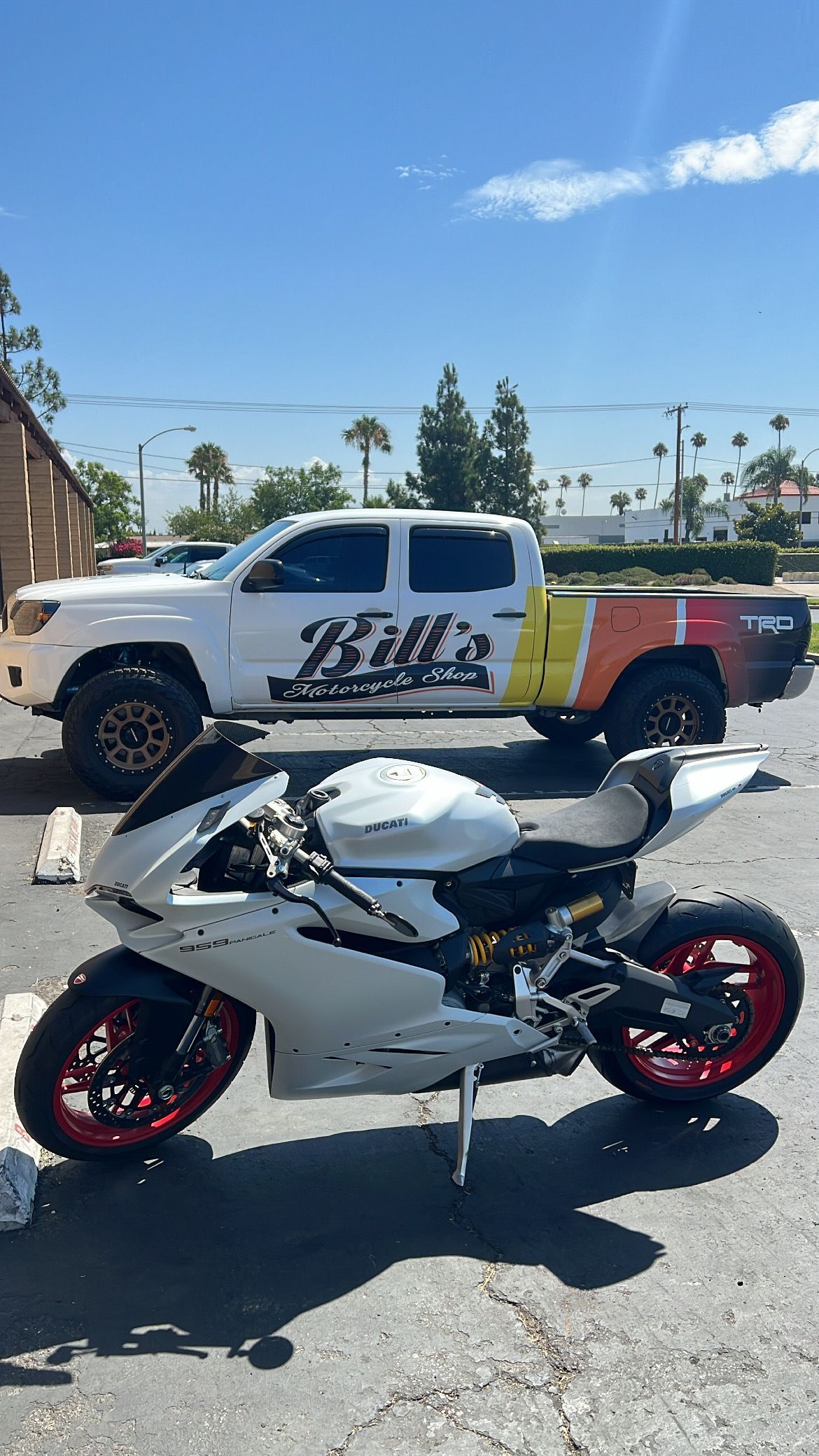 White motorcycle parked next to a truck with colored wrap, sunny outdoor setting.