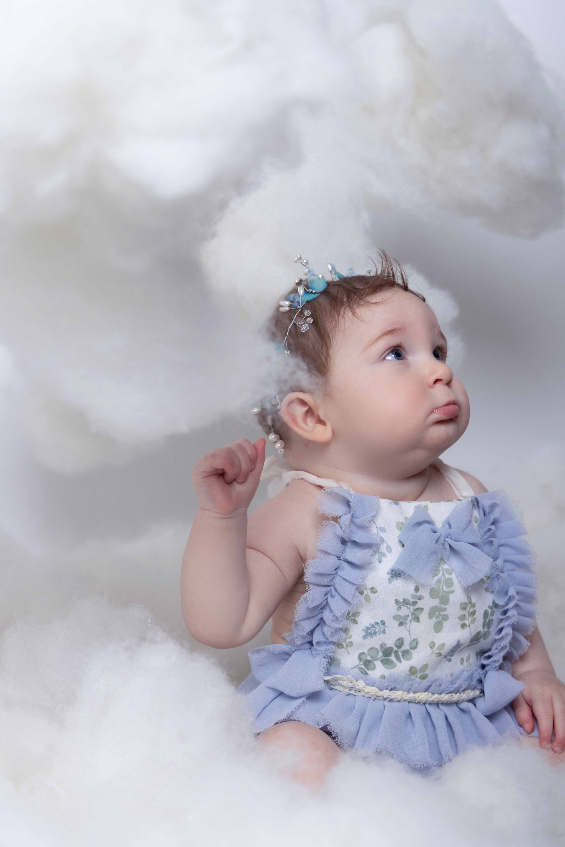 A baby girl in a blue dress is sitting in the clouds.