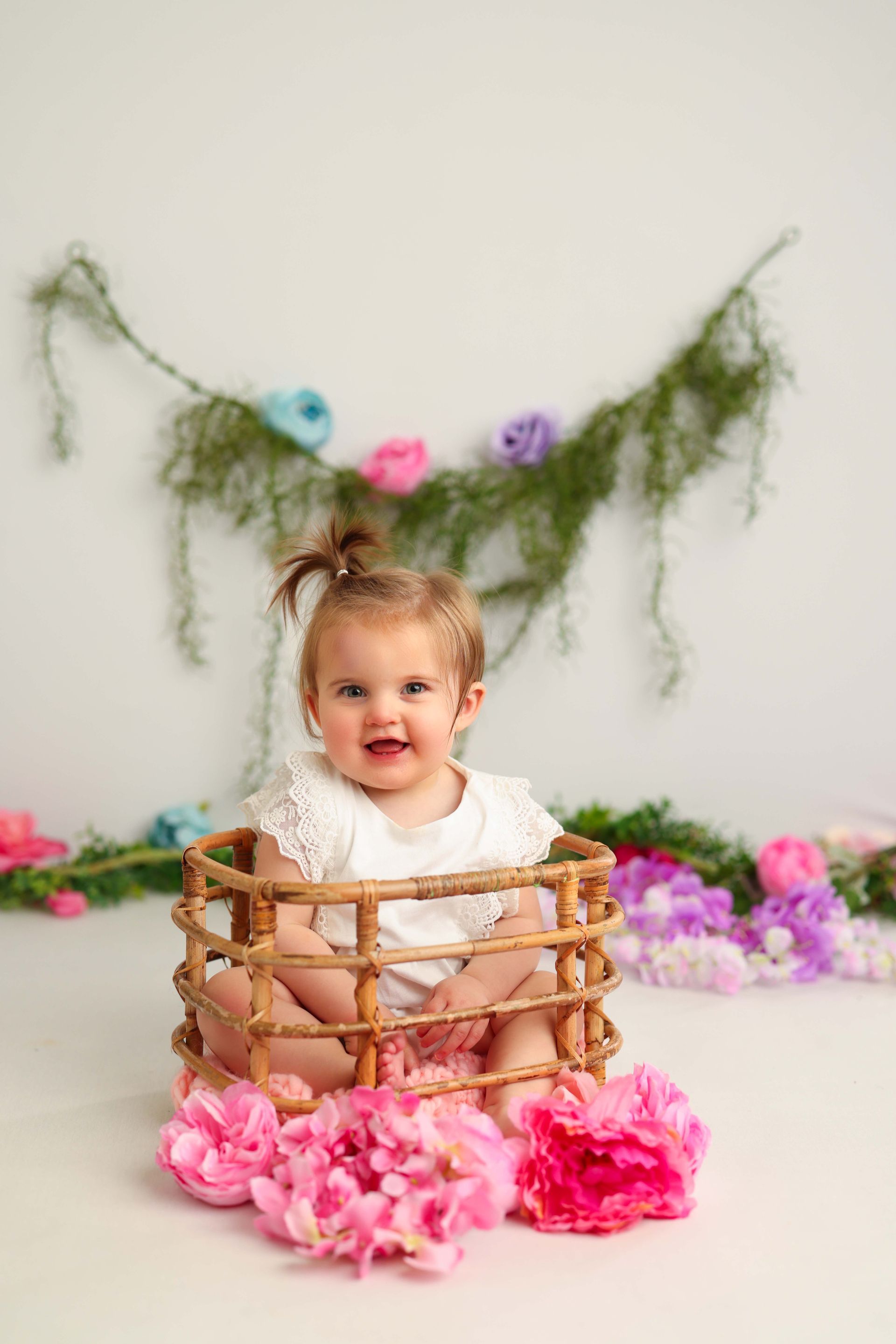 A baby girl is sitting in a basket surrounded by pink flowers.