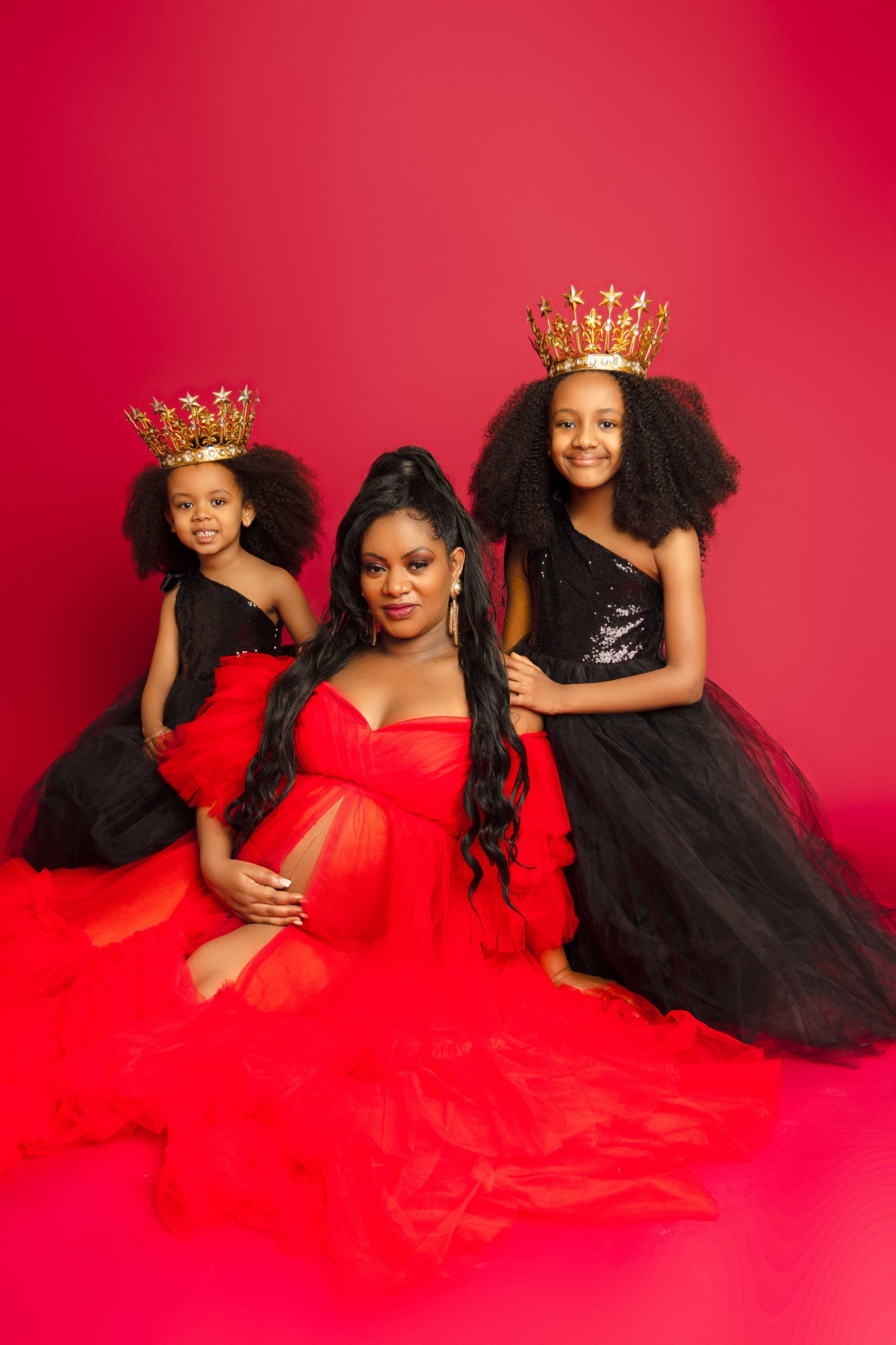 A pregnant woman is sitting next to two little girls wearing crowns.