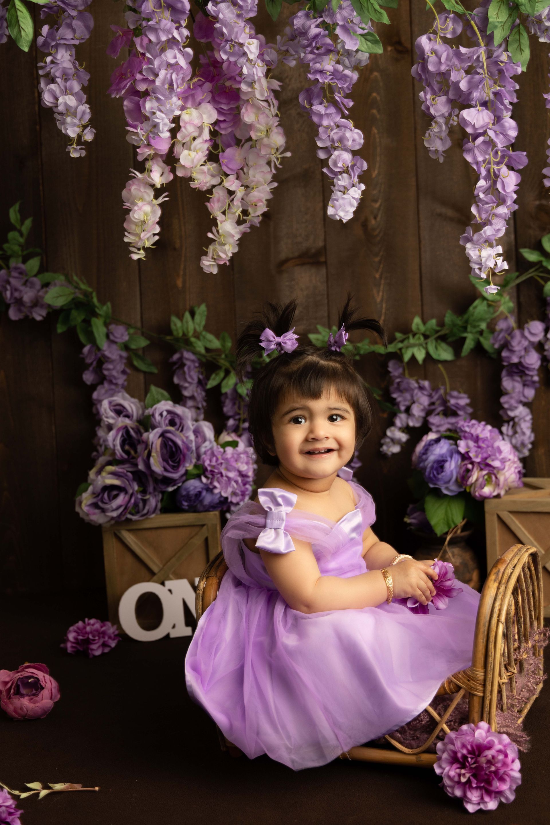 A little girl in a purple dress is sitting in a basket surrounded by purple flowers.