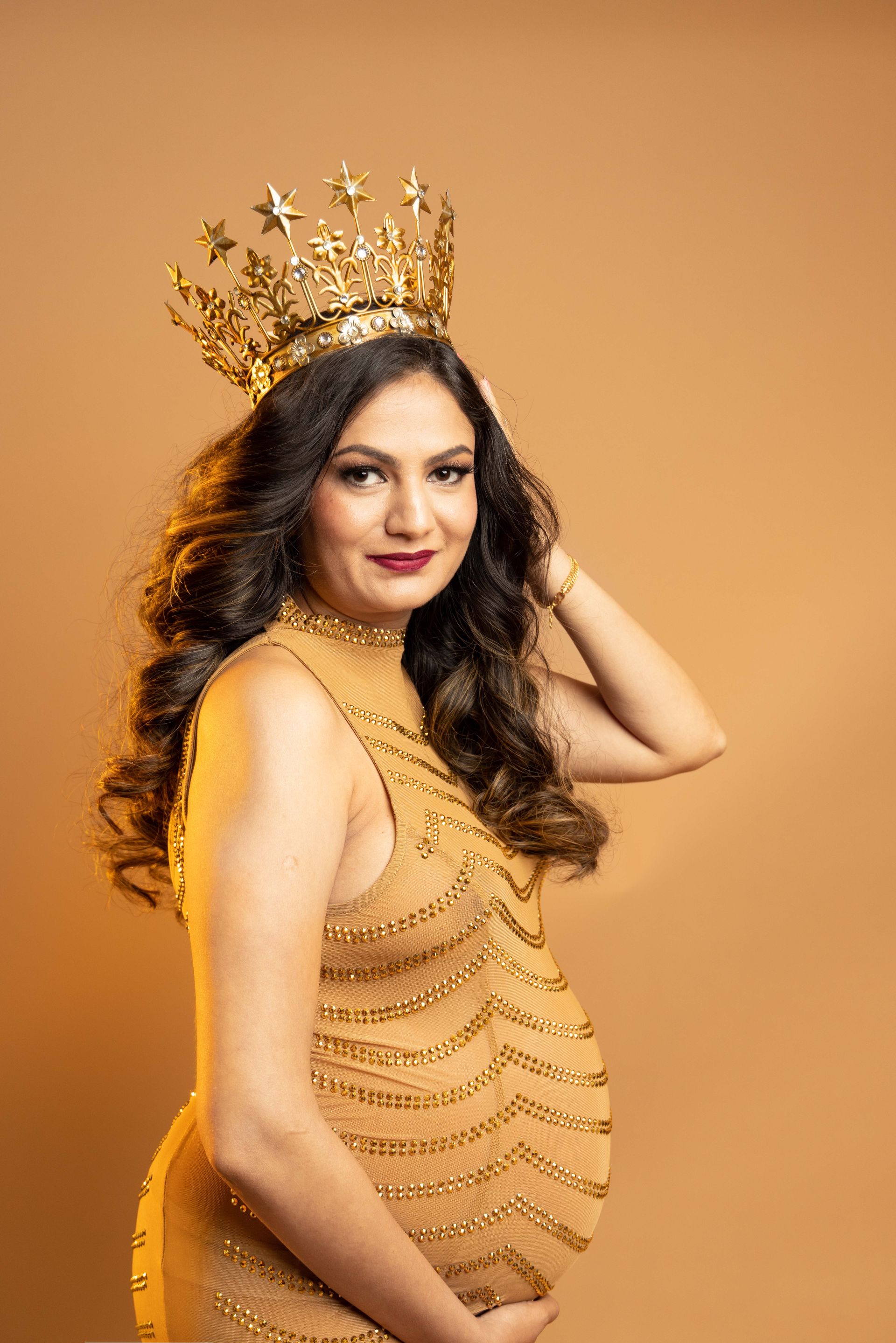 A pregnant woman wearing a crown and a gold dress is holding her belly.