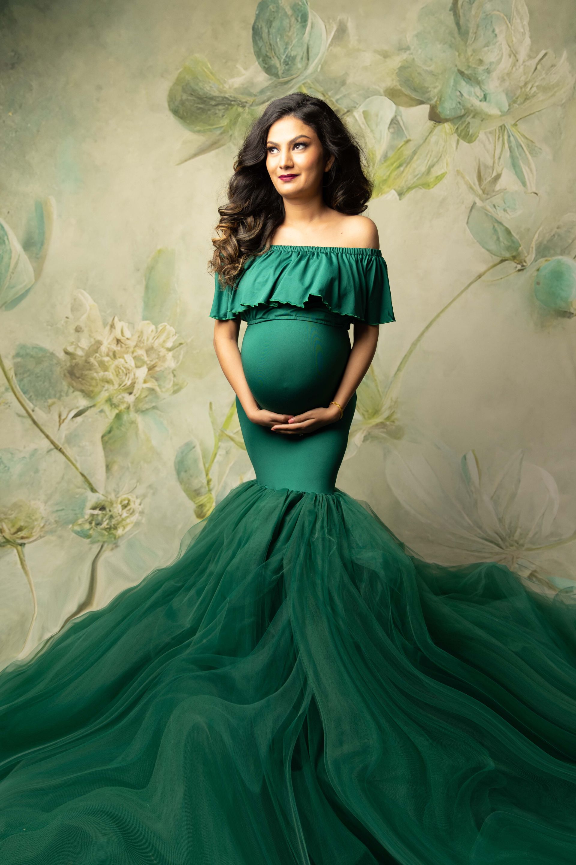A pregnant woman in a green dress is standing in front of a floral background.