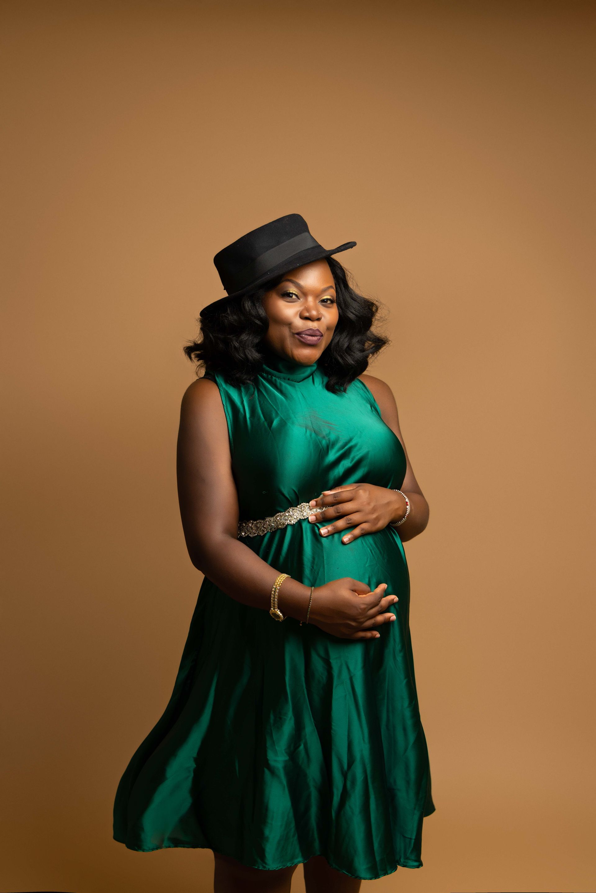 A pregnant woman in a green dress and hat is holding her belly.