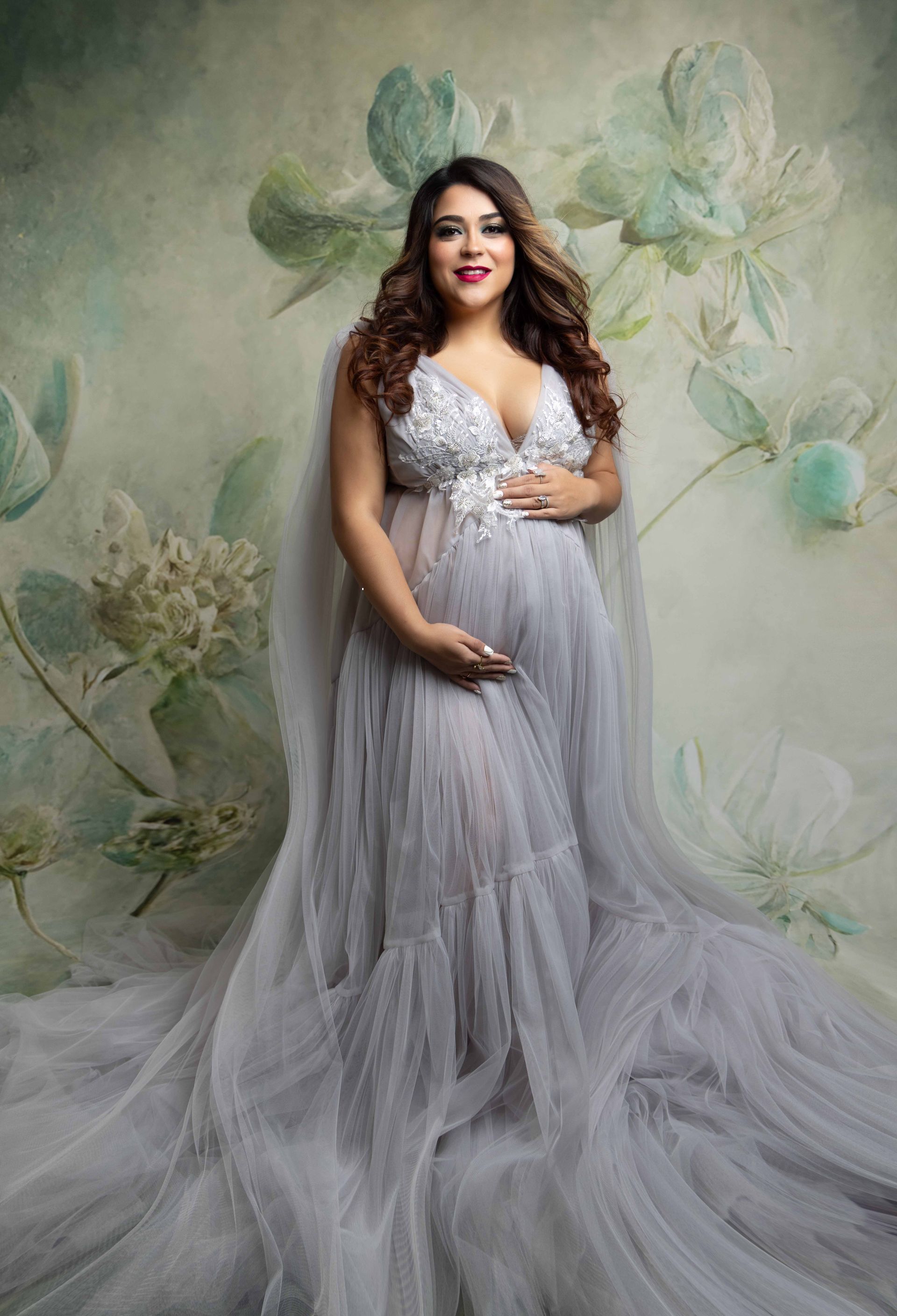 A pregnant woman in a long gray dress is standing in front of a floral background.
