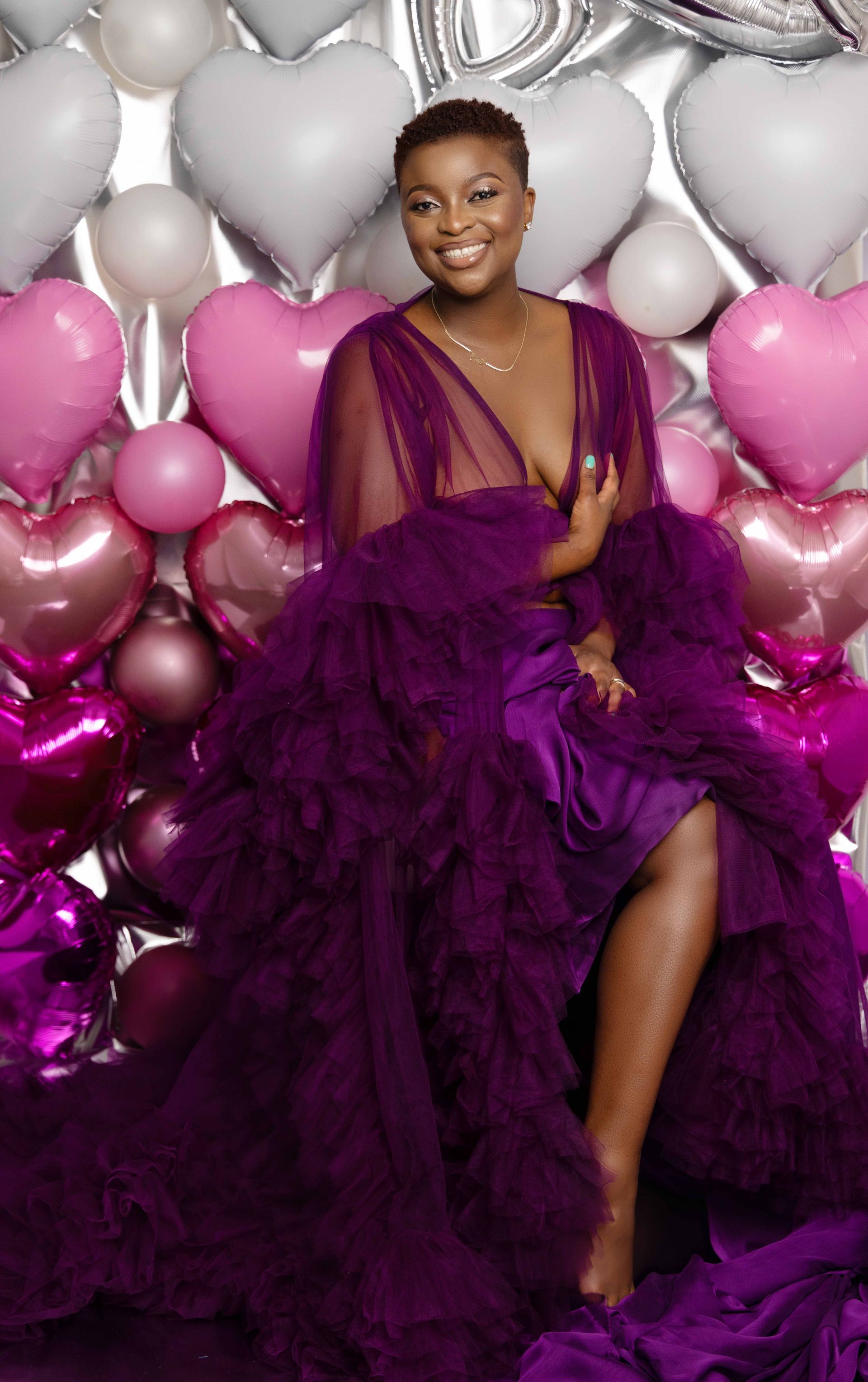 A woman in a purple dress is sitting in front of a wall of heart shaped balloons.