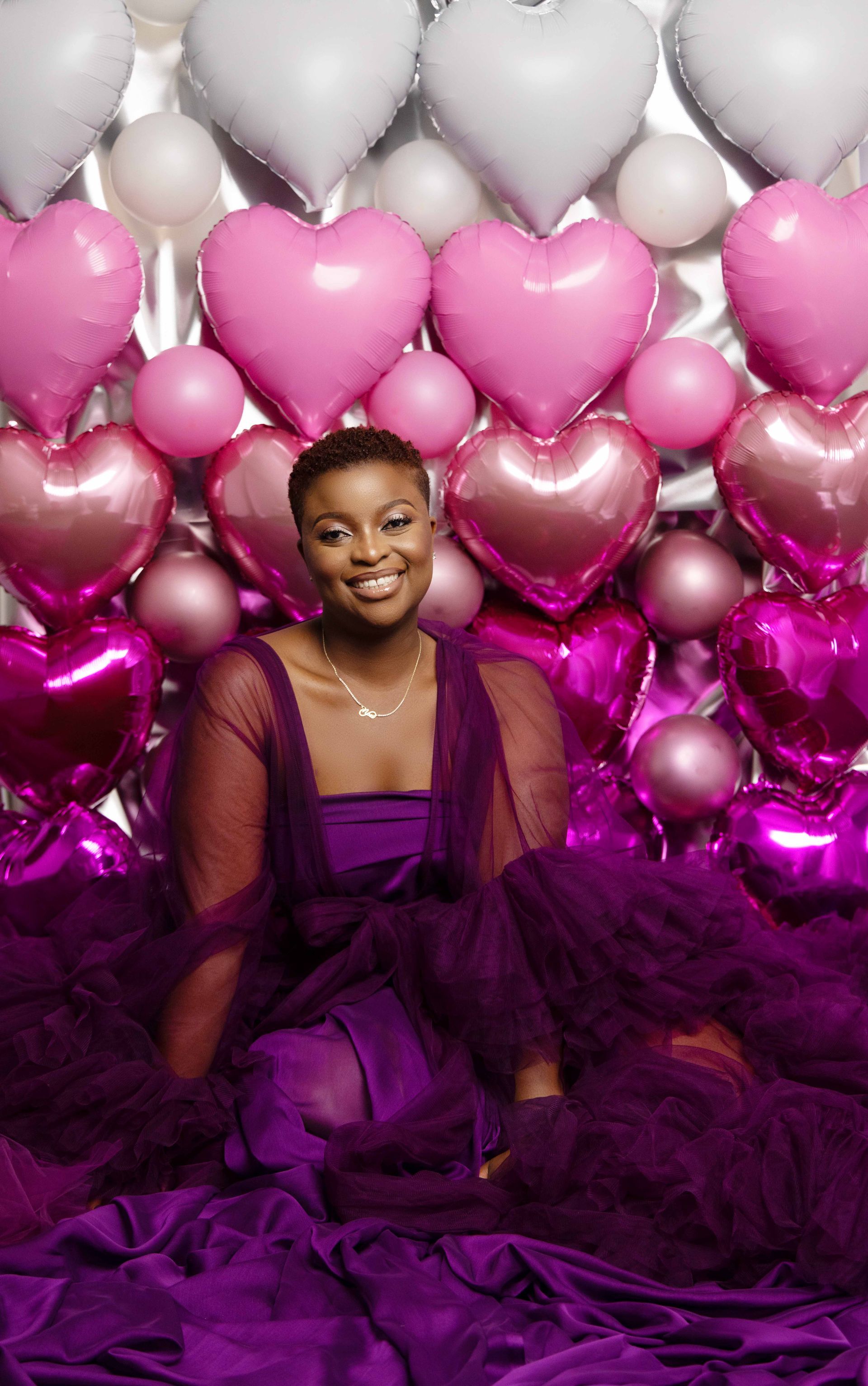 A woman in a purple dress is sitting in front of a wall of heart shaped balloons.