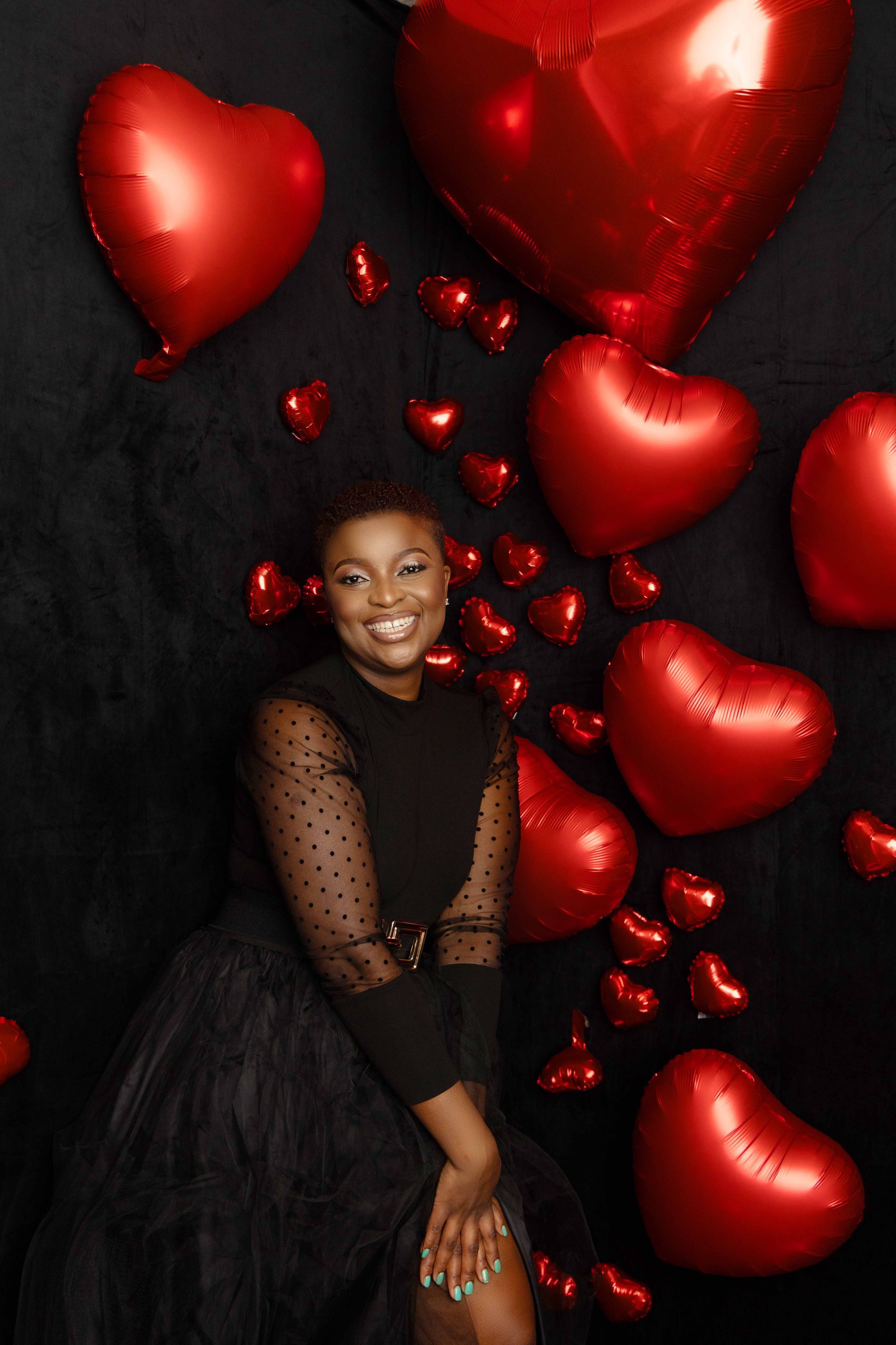 A woman is sitting in front of a wall of red heart shaped balloons.